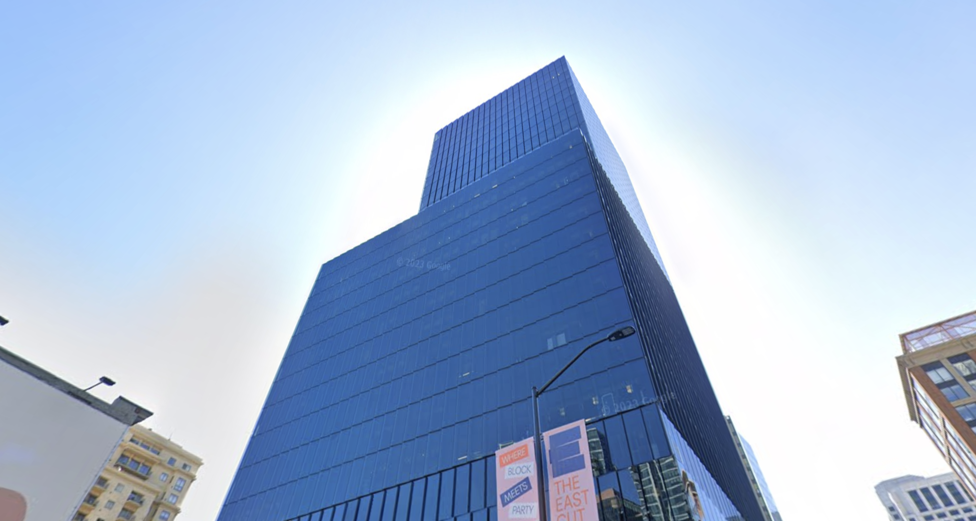 A tall, modern glass skyscraper reflects the sky, surrounded by smaller buildings and banners advertising a block party event.