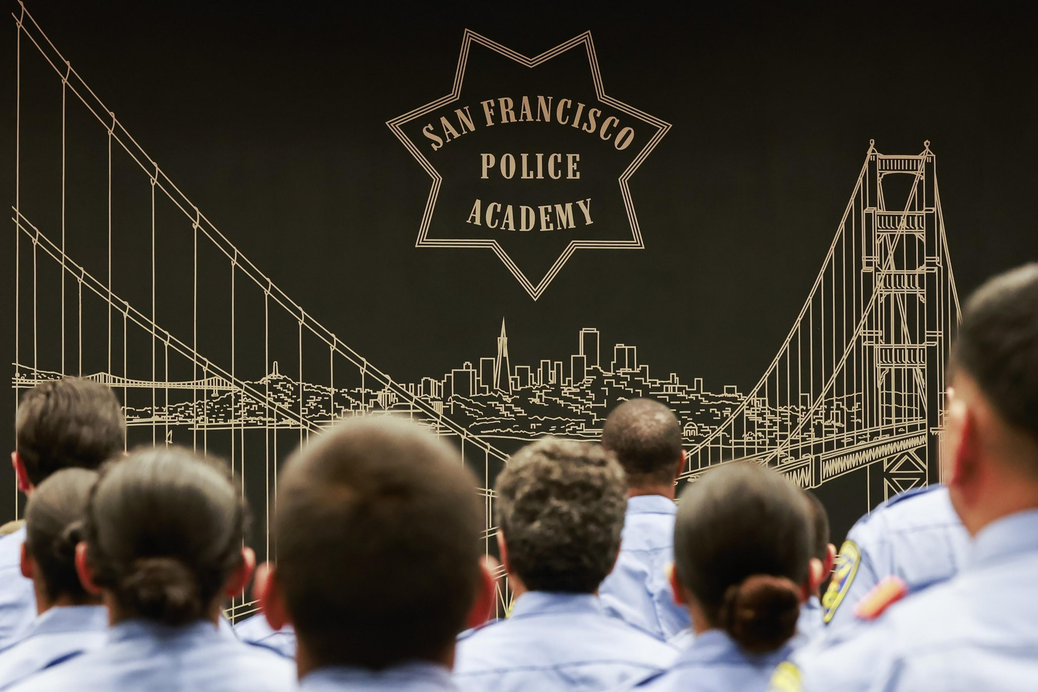 A group of uniformed individuals sits facing a large wall mural of the San Francisco skyline and bridge, with "San Francisco Police Academy" prominently displayed.