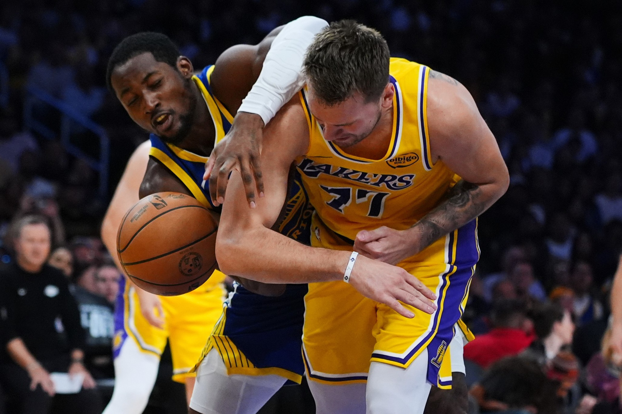 Two basketball players compete intensely for the ball, one in a yellow Lakers jersey and the other in a blue and yellow jersey, with the crowd blurred in the background.