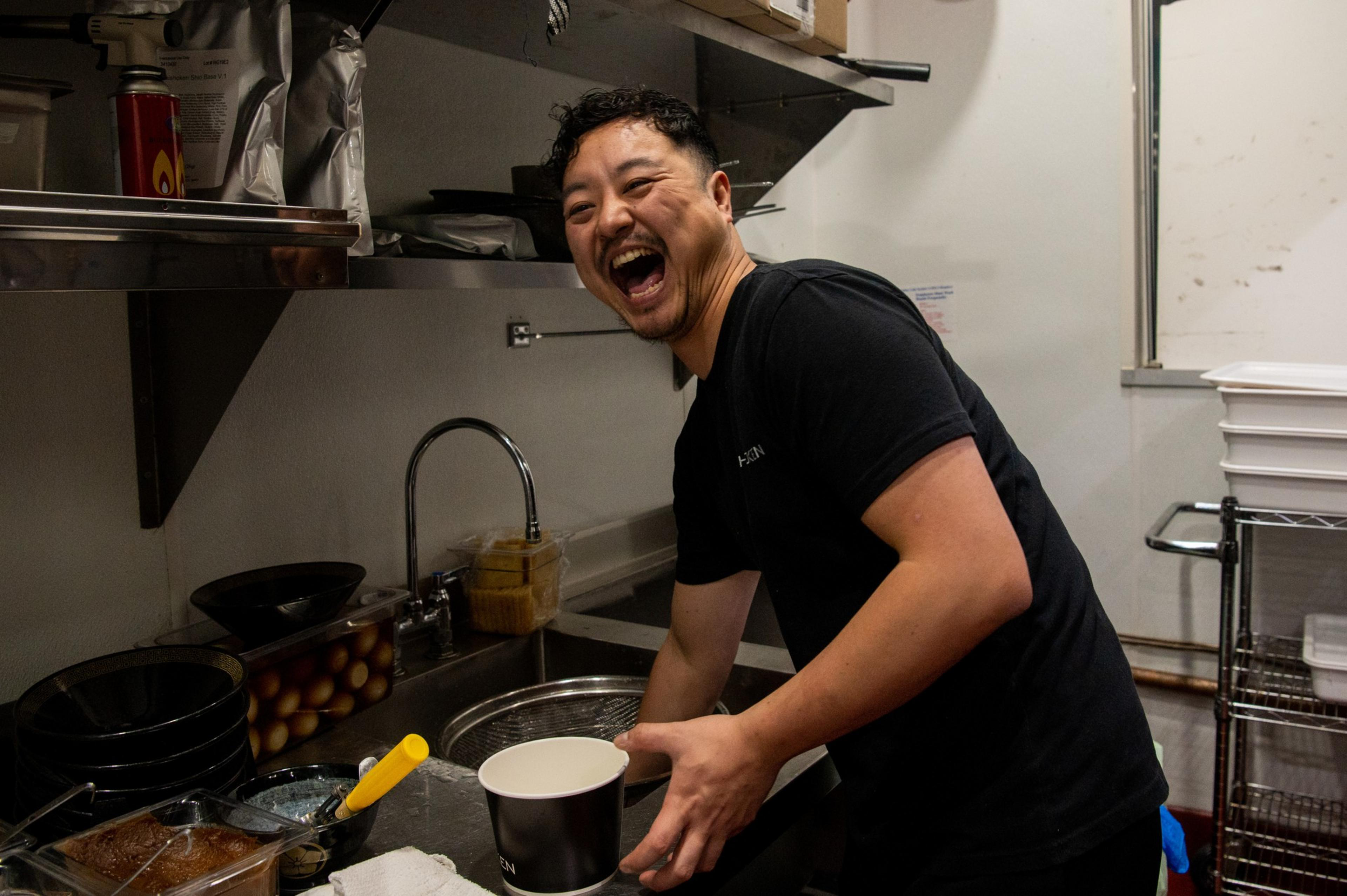 A laughing man stands over a sink in a restaurant kitchen.