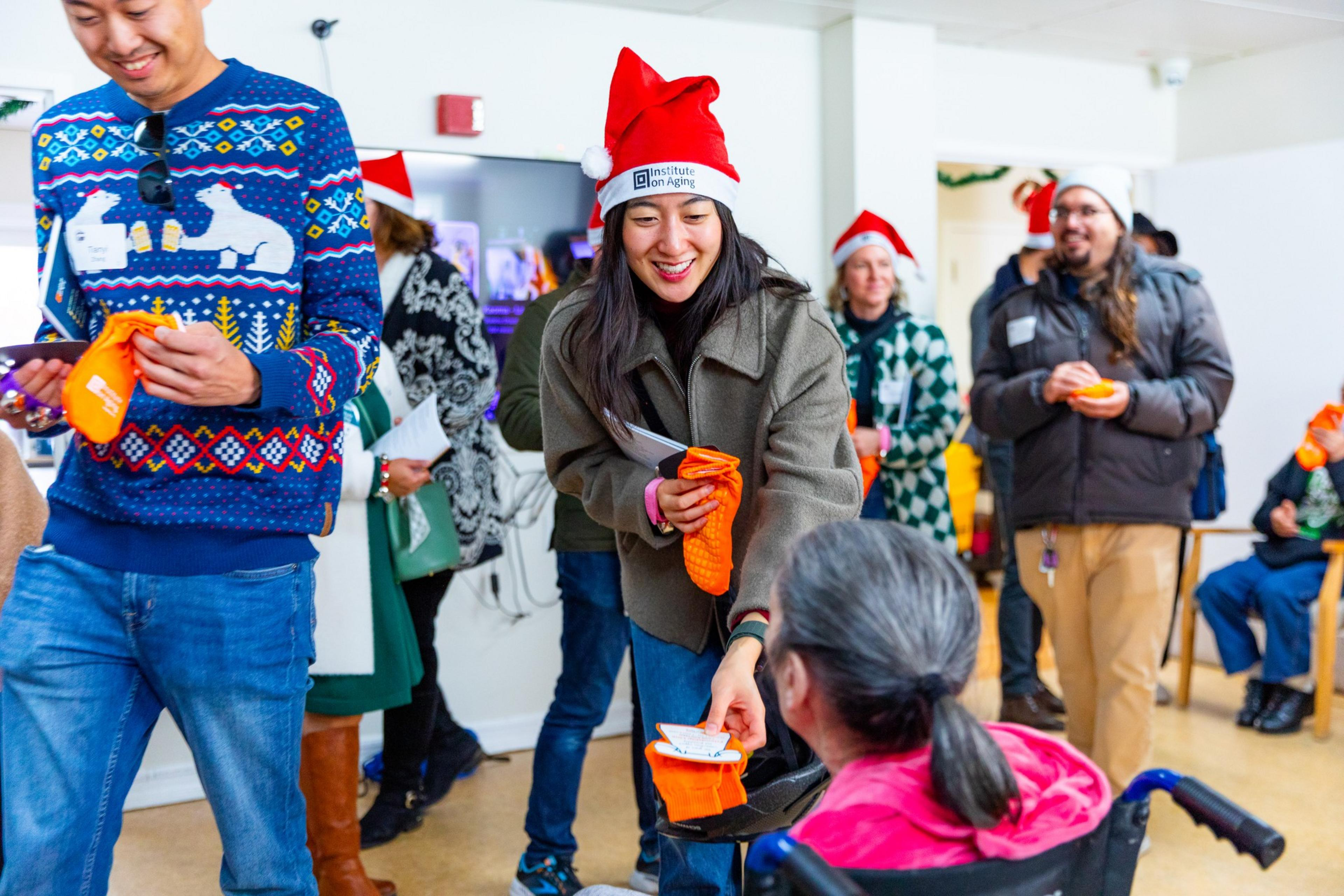 People in festive attire, including Santa hats and bright sweaters, engage in a cheerful gathering, with one person handing an orange item to another in a wheelchair.