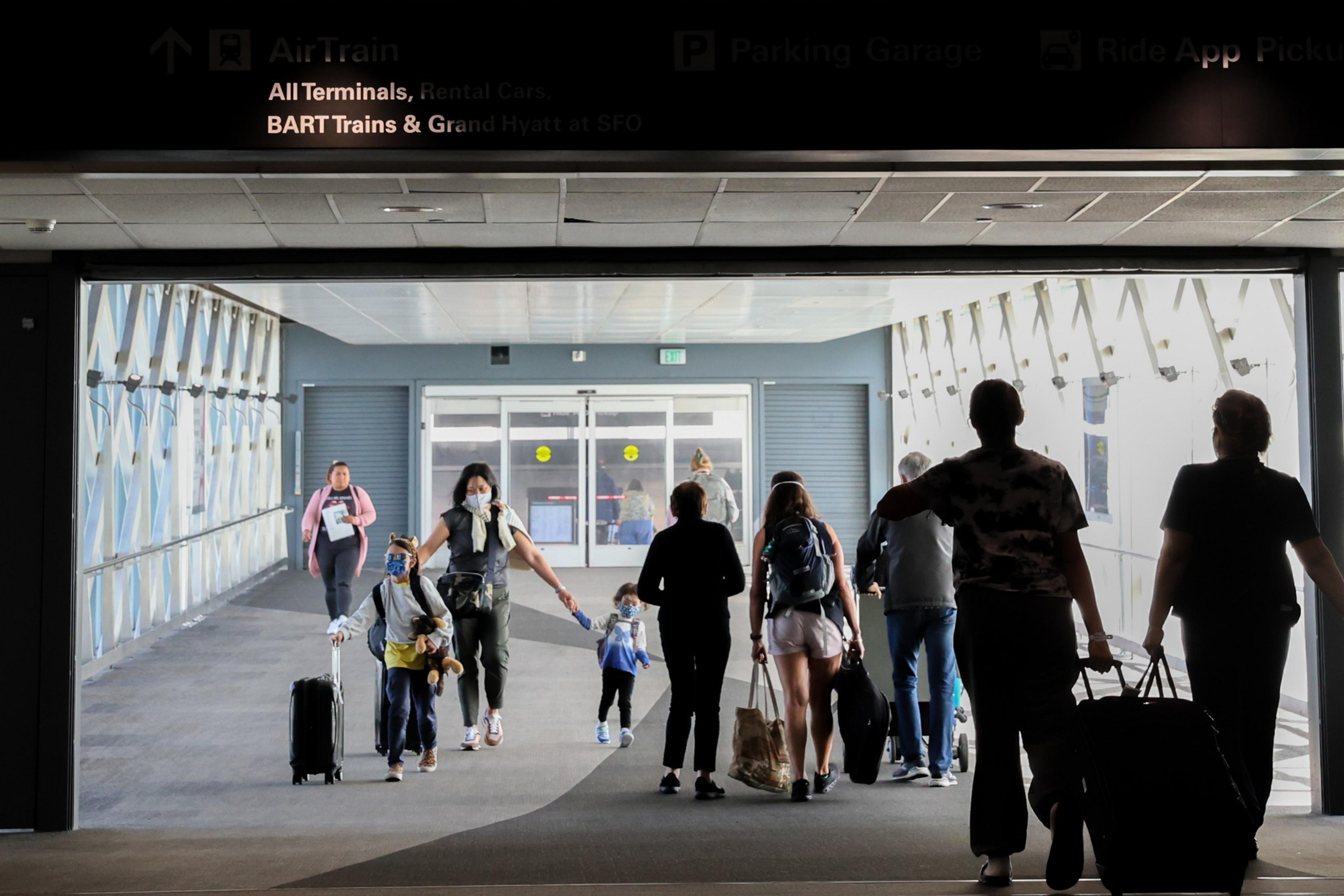 People, some wearing masks, walk through a tunnel with luggage, heading toward doors labeled for terminals, BART trains, and other airport services.