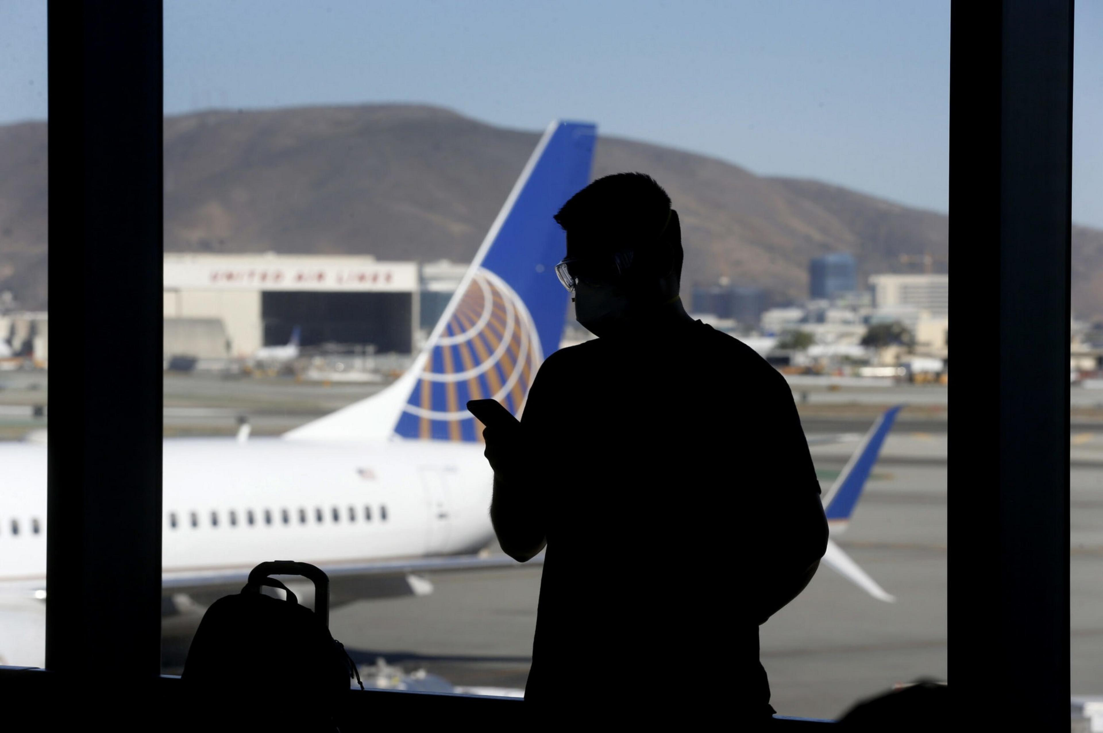 A silhouette of a person holding a phone stands indoors near a window, with a United Airlines plane and hills visible outside.