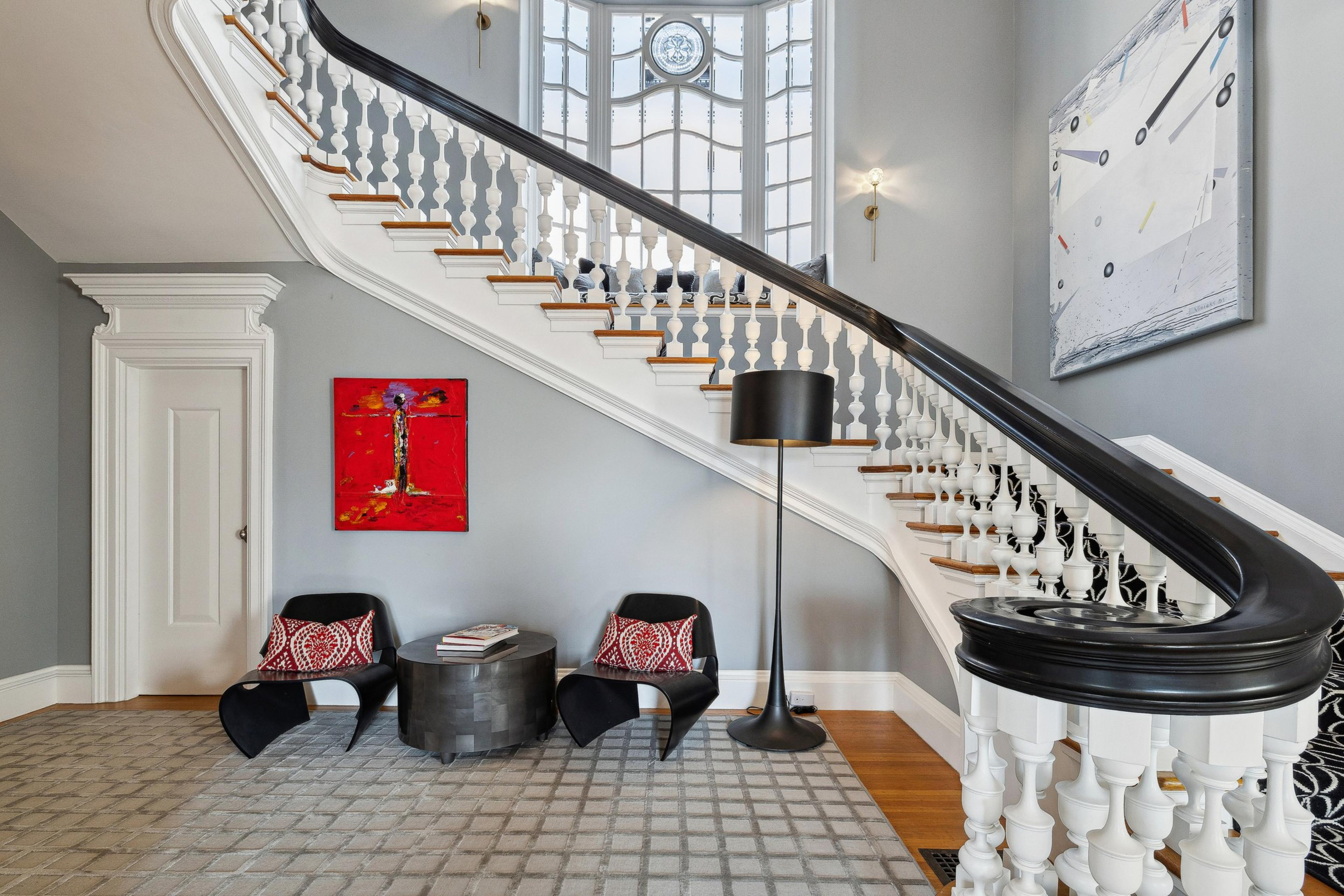 A winding staircase with white spindles and a black handrail leads upstairs in a stylish home. Two modern chairs, a round table, and a tall lamp sit below it.