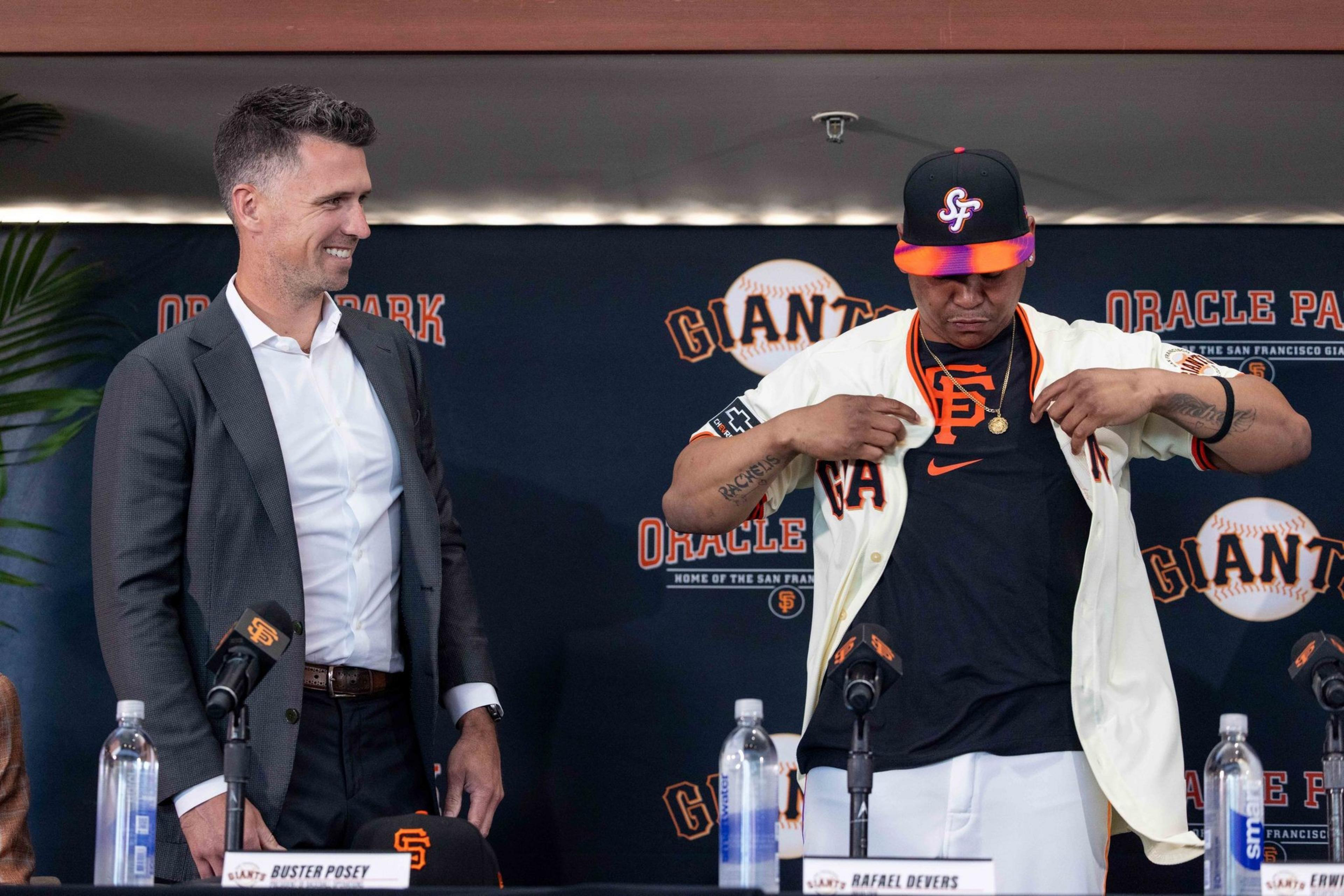 Two people stand at a podium with microphones. One is in a suit smiling, the other wears a baseball jersey and cap, adjusting his shirt. The background says &quot;Giants&quot; and &quot;Oracle Park&quot;.