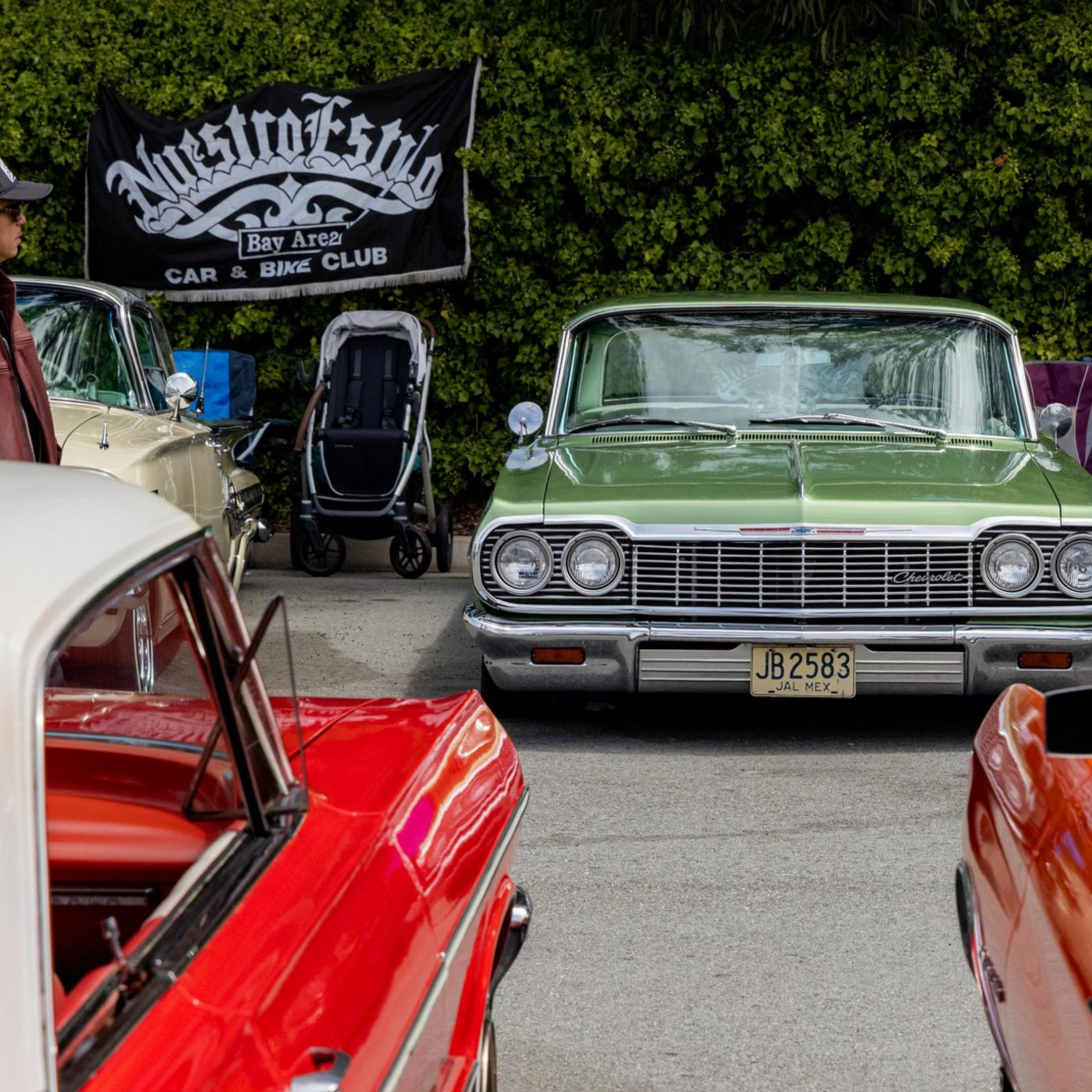 A green vintage car is parked facing forward at a car show, surrounded by other colorful classic cars, with people and a &quot;Maestro Klub&quot; banner in the background.