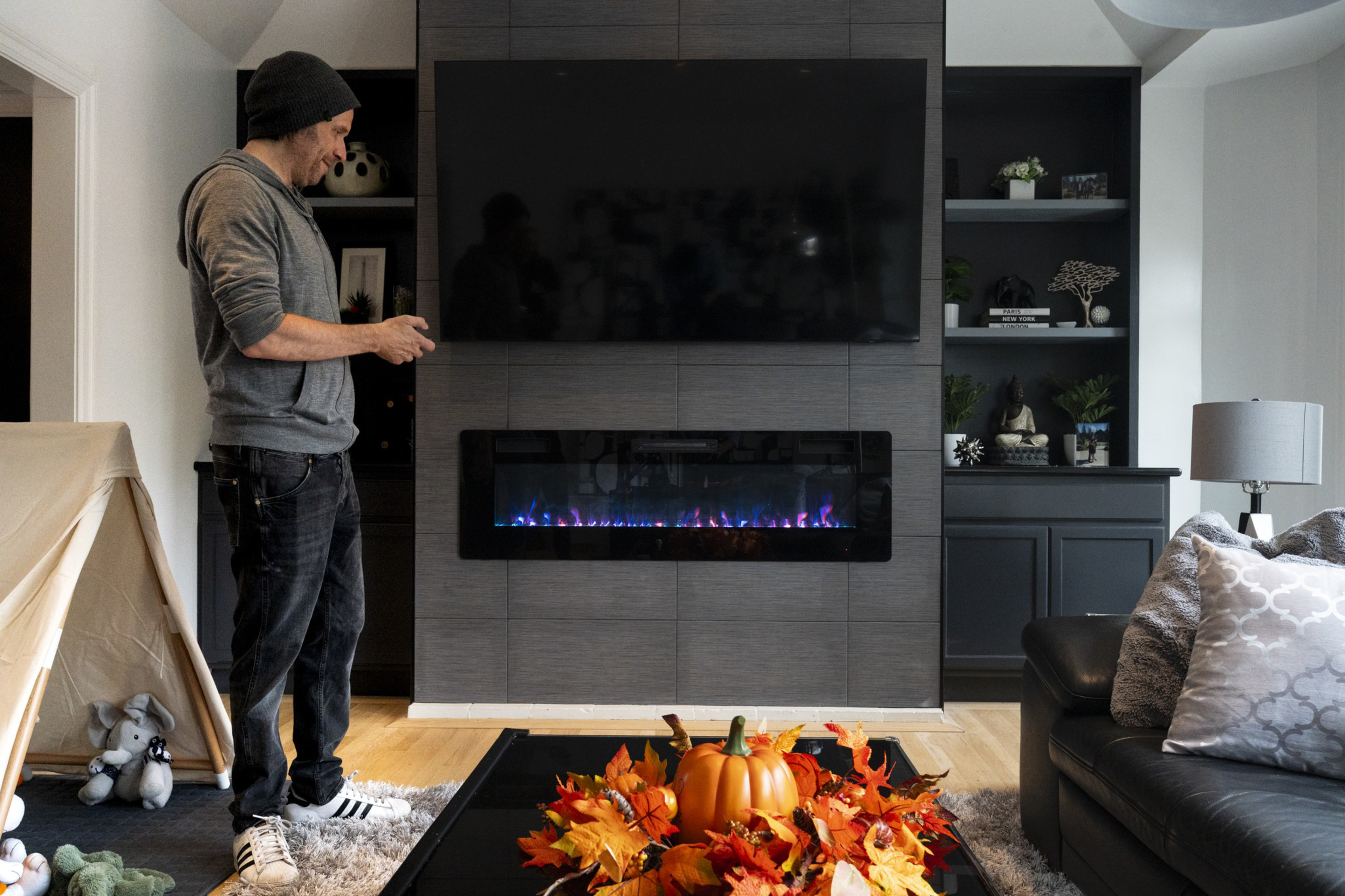 A man in casual clothes stands near a modern electric fireplace under a large TV, with a pumpkin and autumn leaves centerpiece on the table.