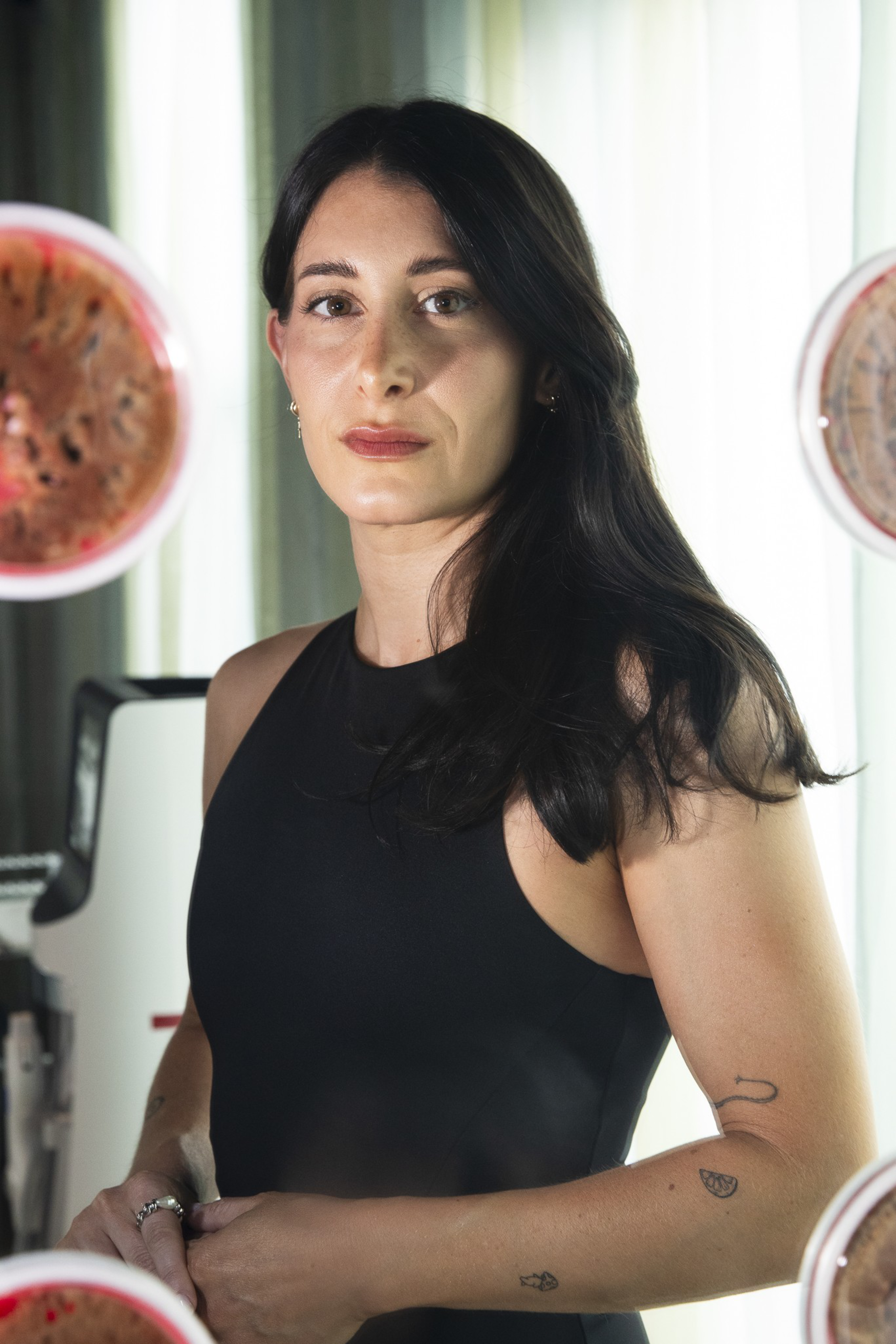 A woman with long dark hair wears a black sleeveless top, standing confidently with subtle tattoos on her arms and soft natural lighting behind her.