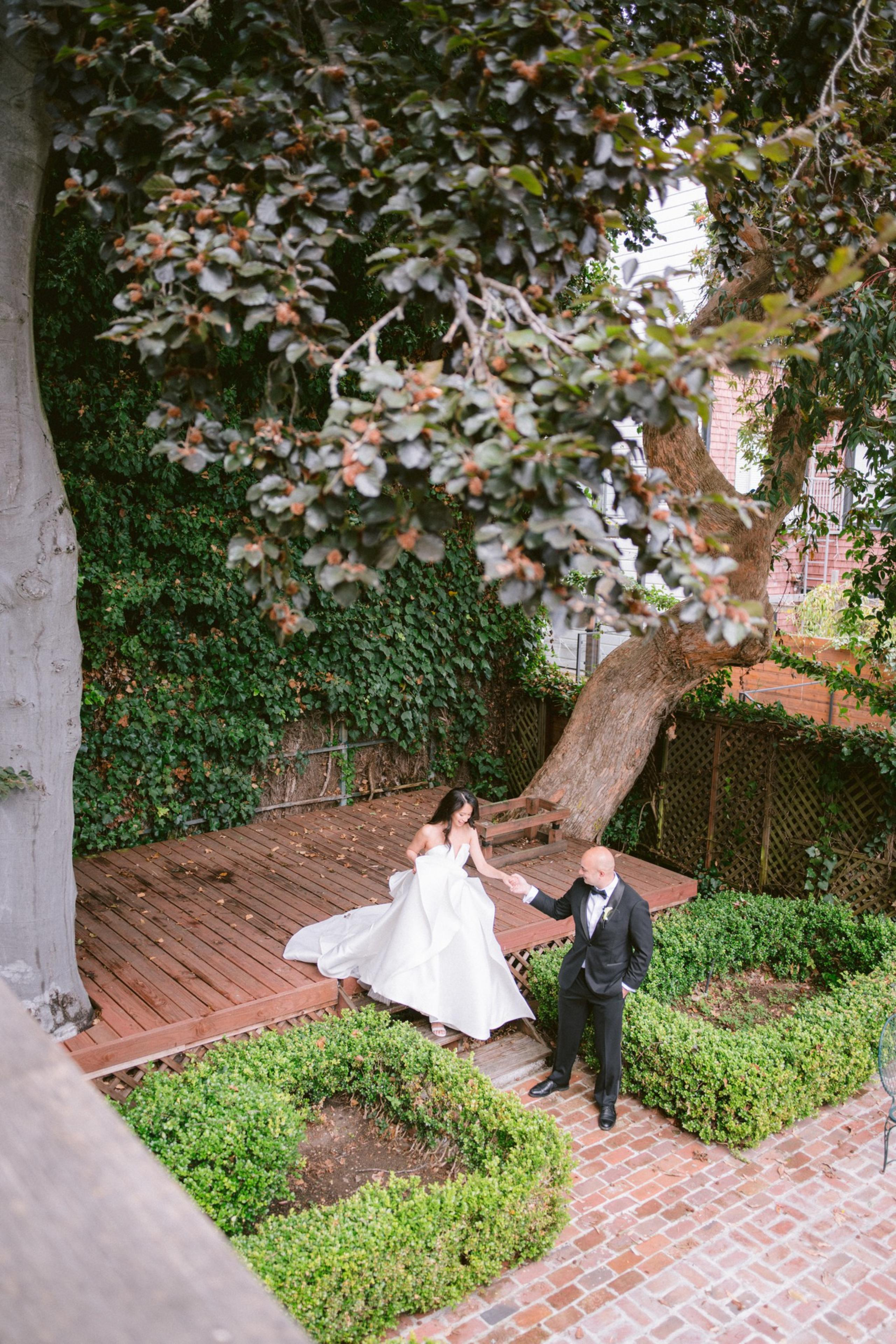 A bride in a white gown steps down from a wooden deck, holding the hand of a groom in a black suit, surrounded by green bushes and large trees.