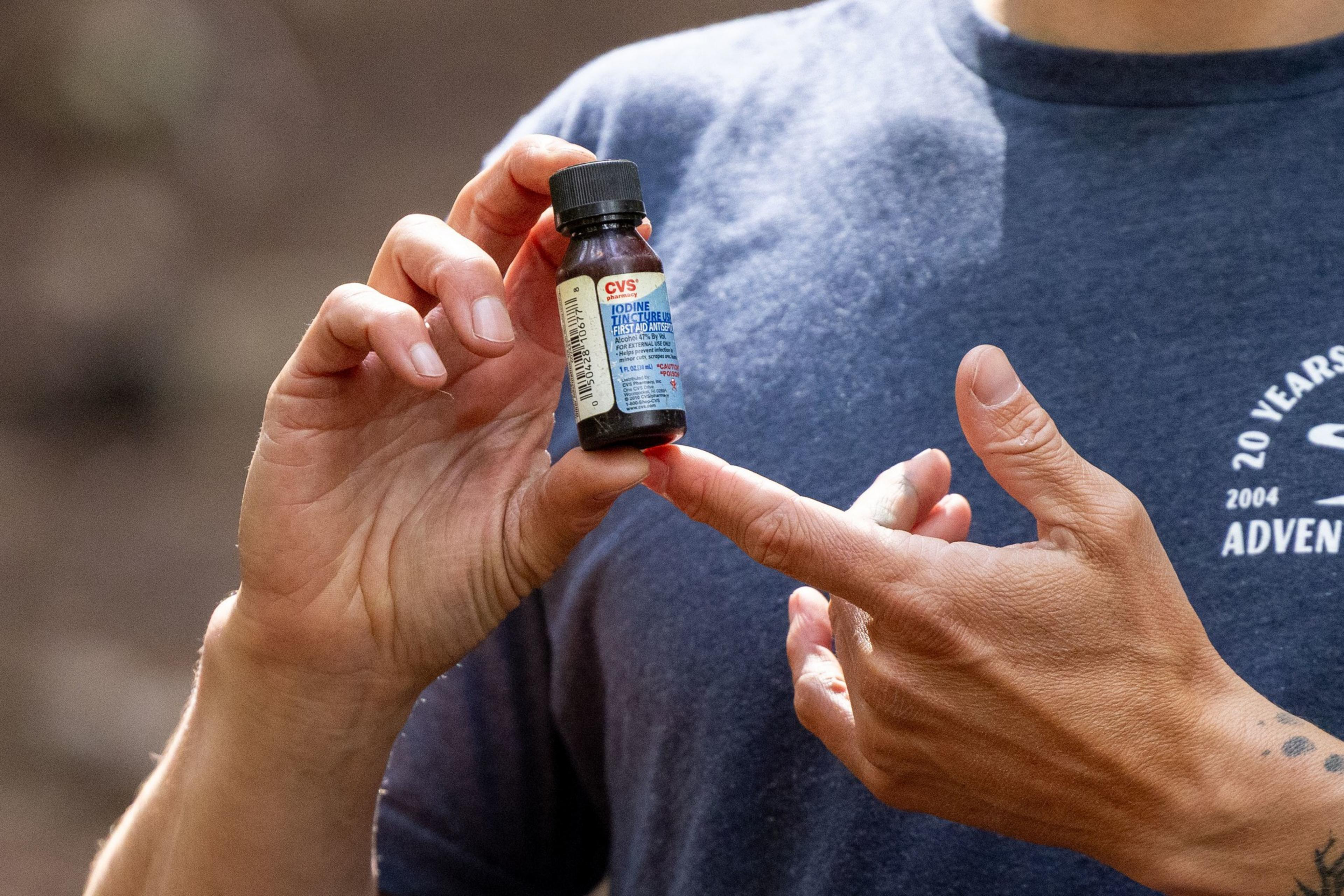 A person is holding a small bottle of CVS iodine tincture with one hand for stability and pointing at it with the other, against a blurred background.
