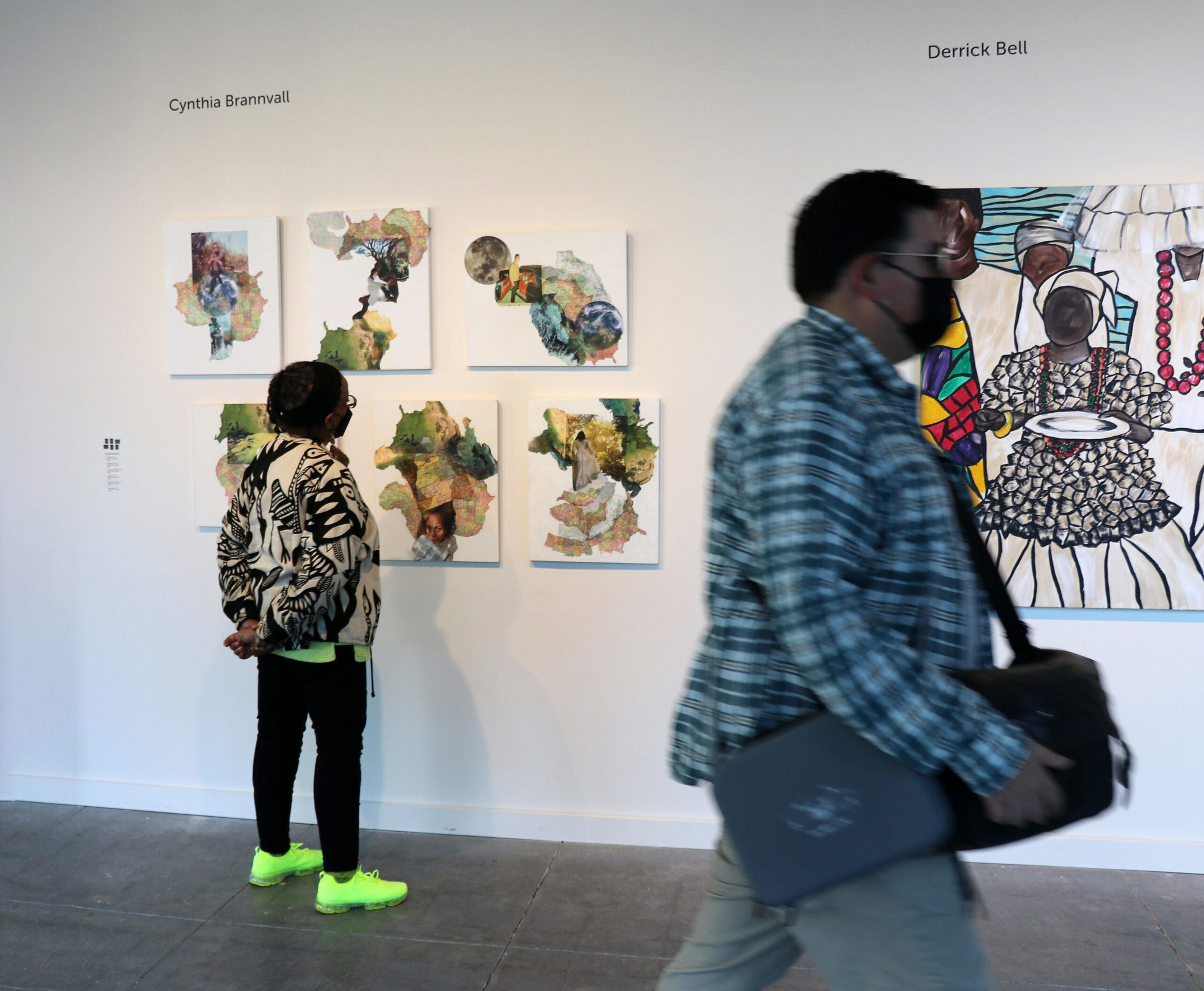A person in bright yellow shoes and patterned jacket observes six abstract paintings, while another person wearing a mask and plaid jacket walks past.