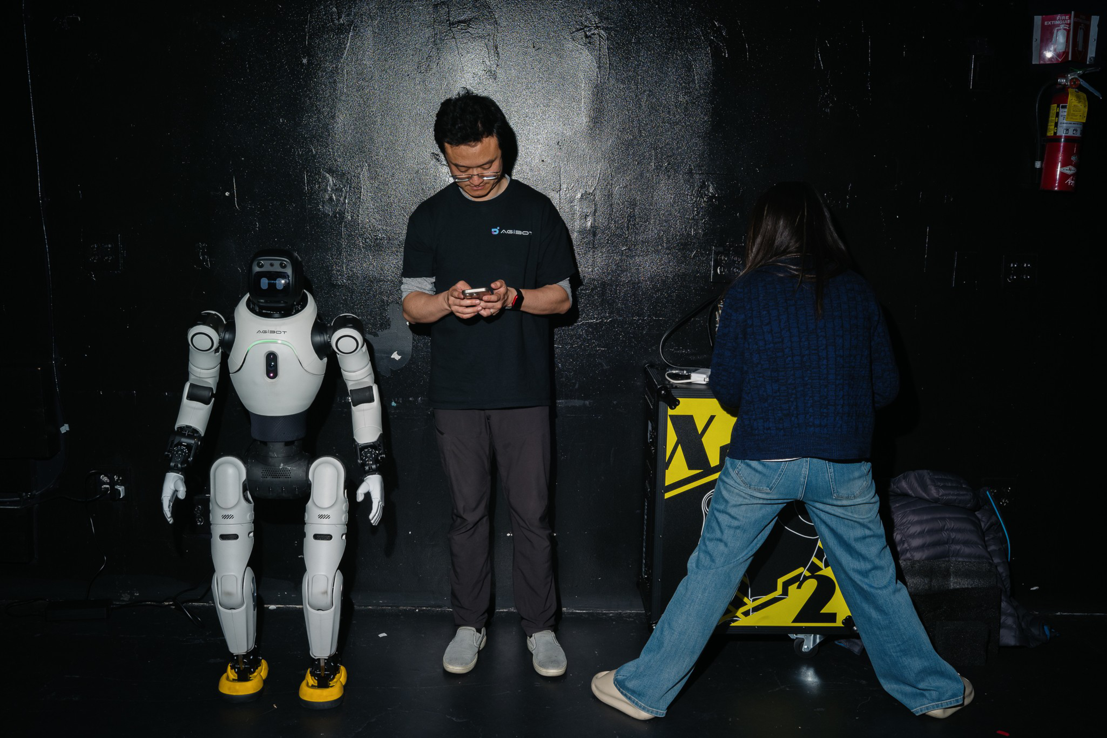 A humanoid robot stands next to a man looking at his phone, while a woman in a dark sweater and jeans faces a black and yellow box with her legs wide apart.