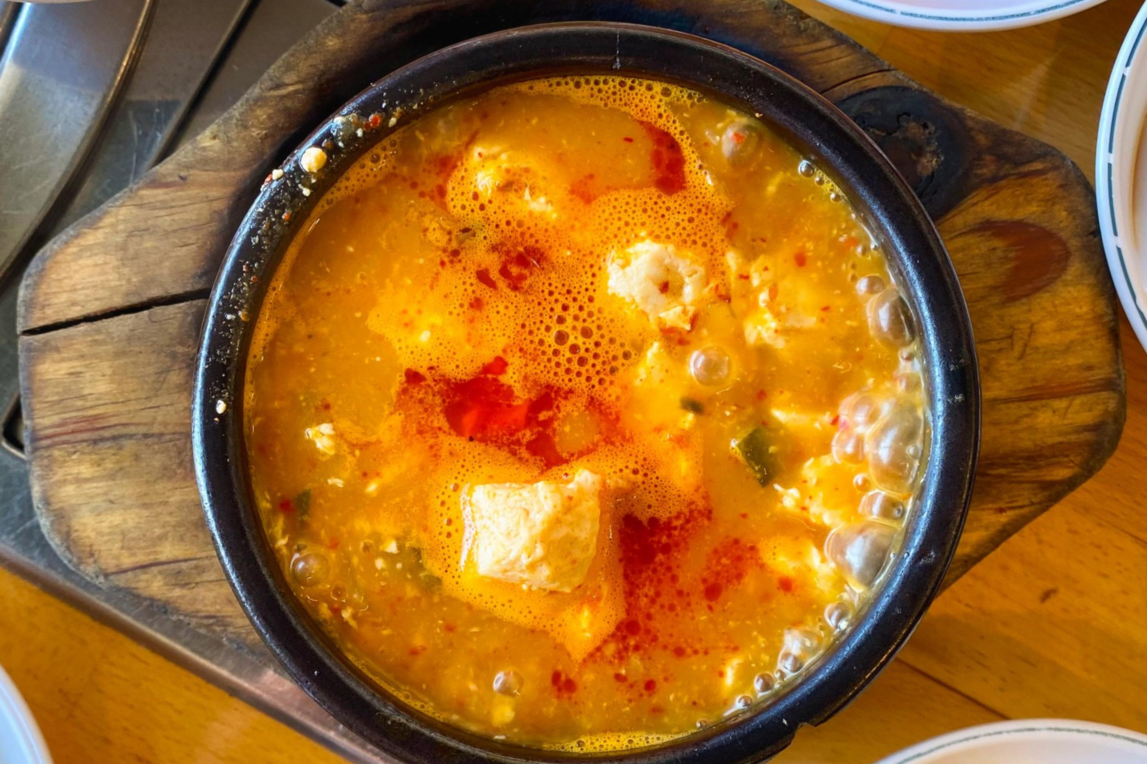 A bubbling pot of stew is surrounded by small bowls containing side dishes like kimchi, radish cubes in red sauce, and bean sprouts on a wooden table.