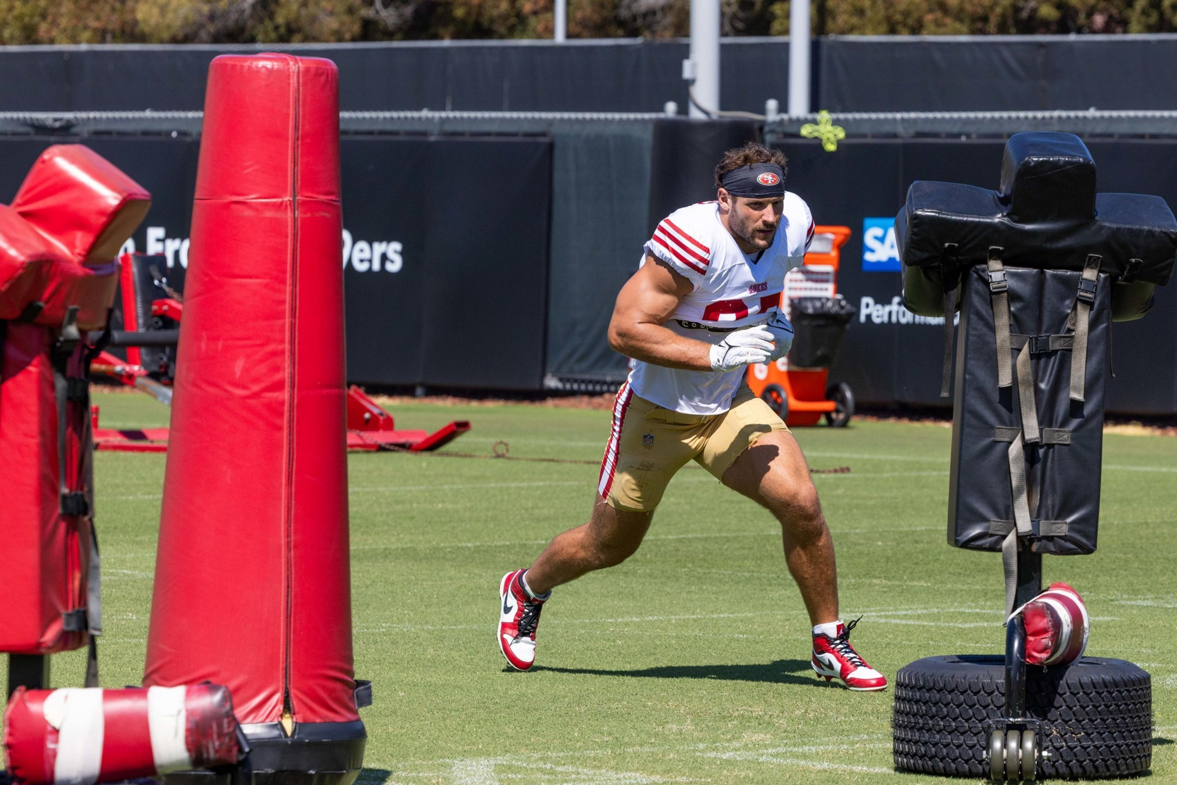 A football player in a white and red jersey and gold pants runs on grass between large red and black training dummies during practice.