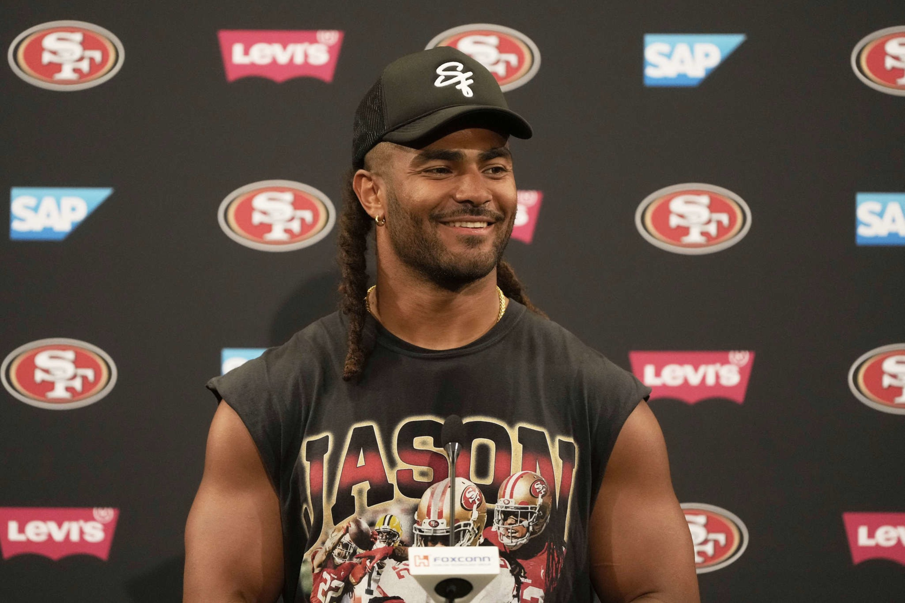 A person with long hair smiles at a press conference, wearing a black cap and a sleeveless shirt featuring football players. The backdrop has SAP and Levi’s logos.