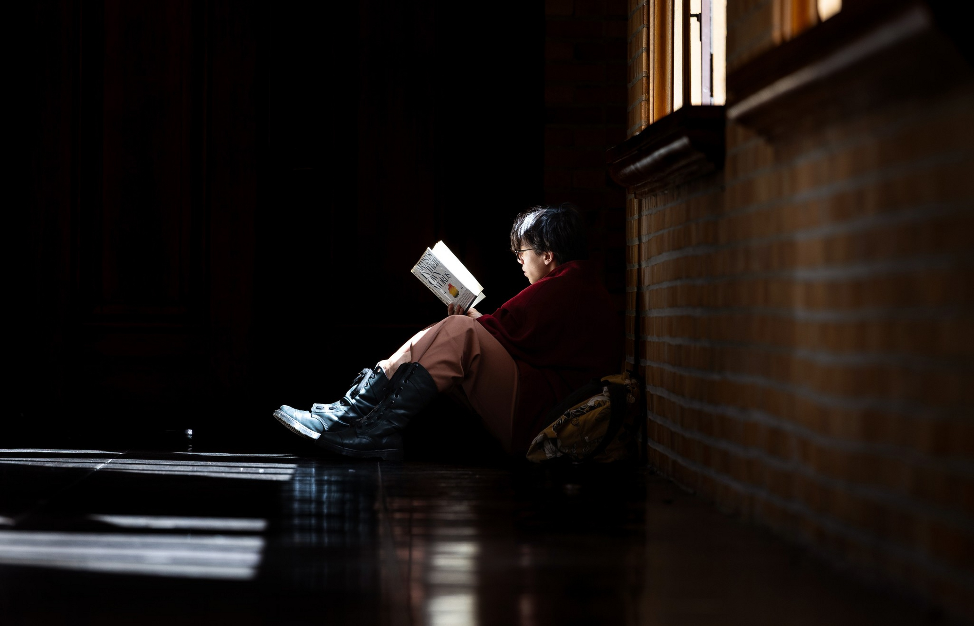 A person with short hair sits on the floor by a window, reading a book, illuminated by natural light in an otherwise dark room.