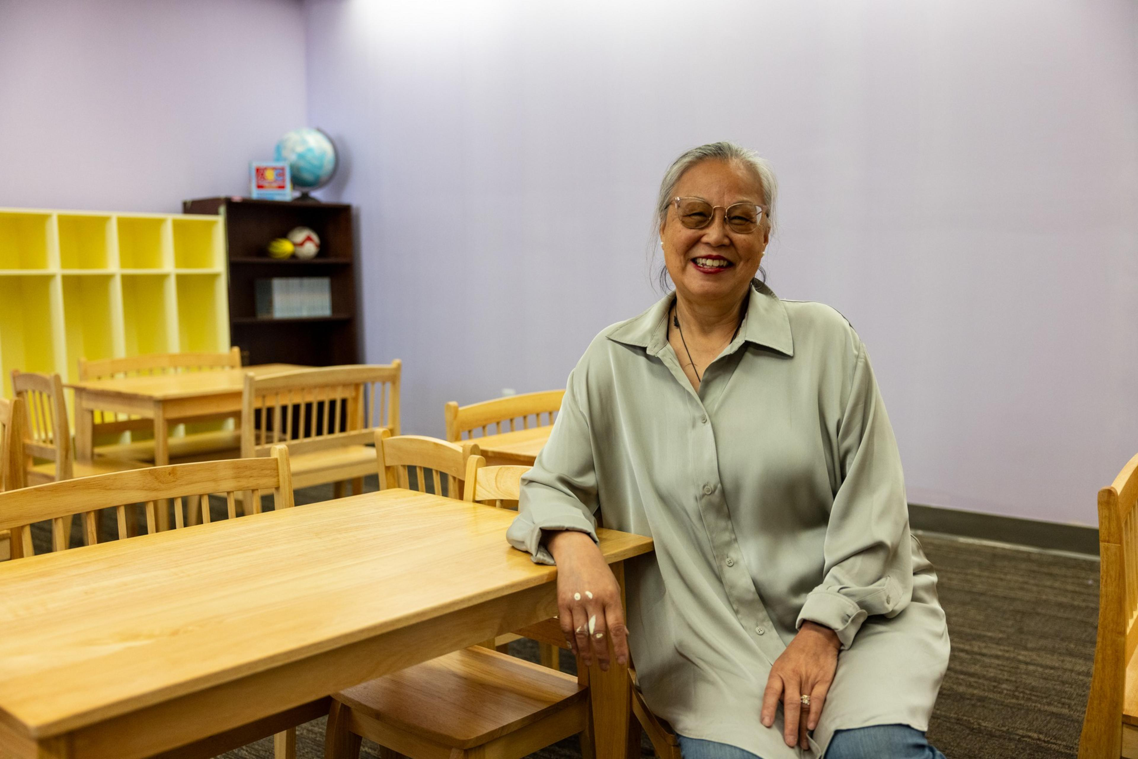 An older woman with glasses smiles while sitting on a chair in a classroom with wooden tables, chairs, a globe, and shelves in the background.