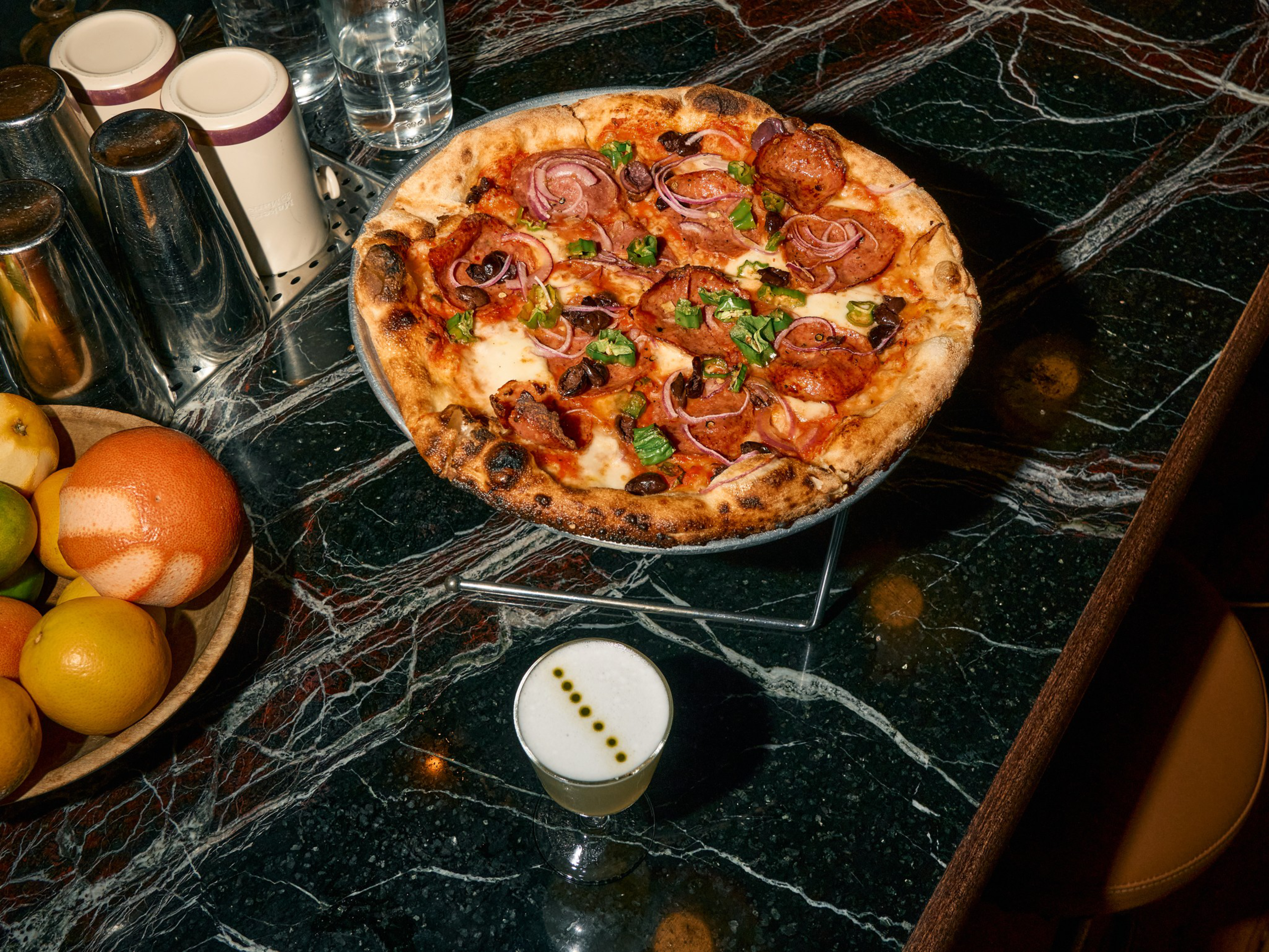 A pepperoni pizza topped with red onions, olives, and green herbs sits on a stand, next to a creamy cocktail with five drops of bitters on top.