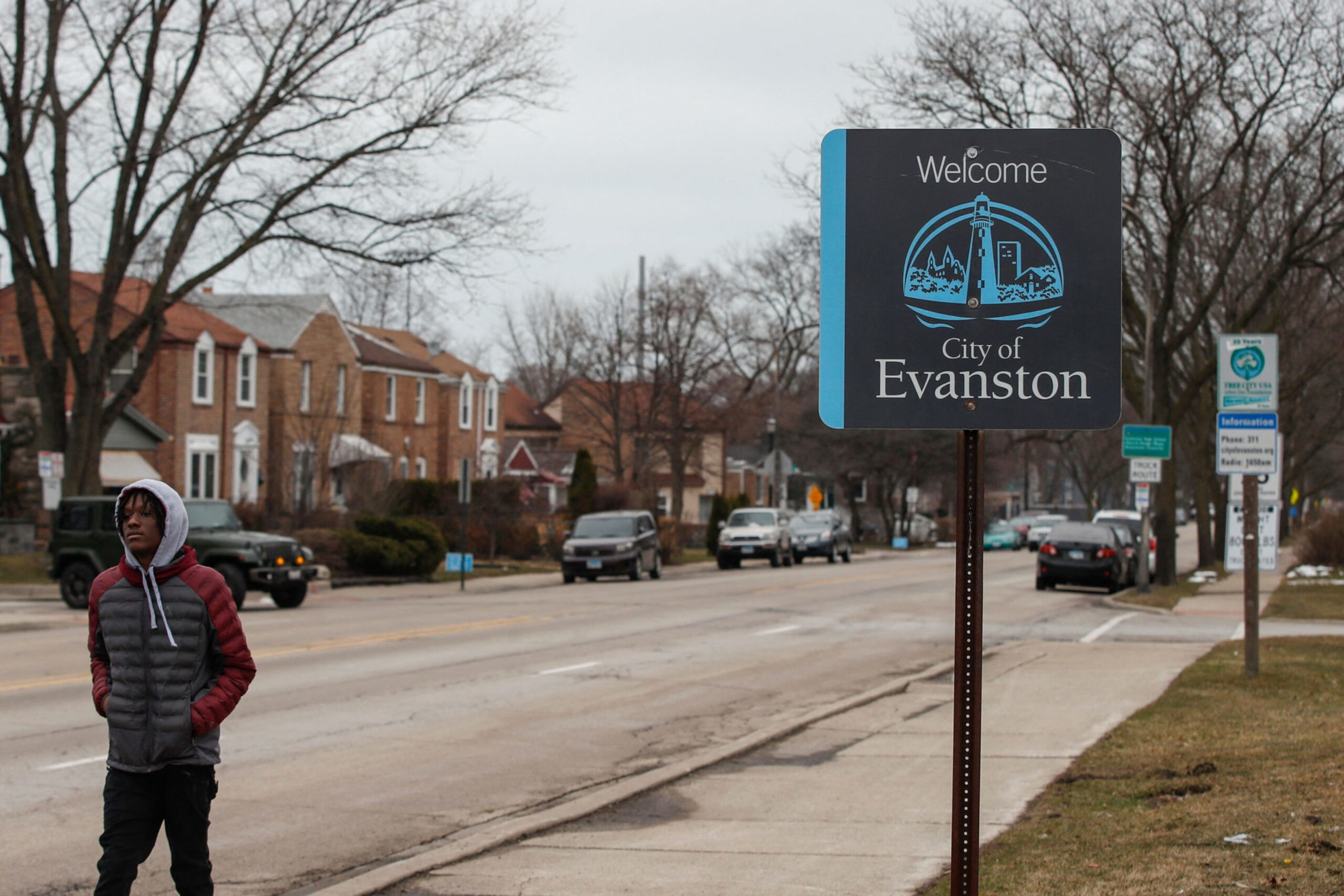 A person in a hooded jacket walks along a street lined with bare trees and brick houses next to a "Welcome City of Evanston" sign.
