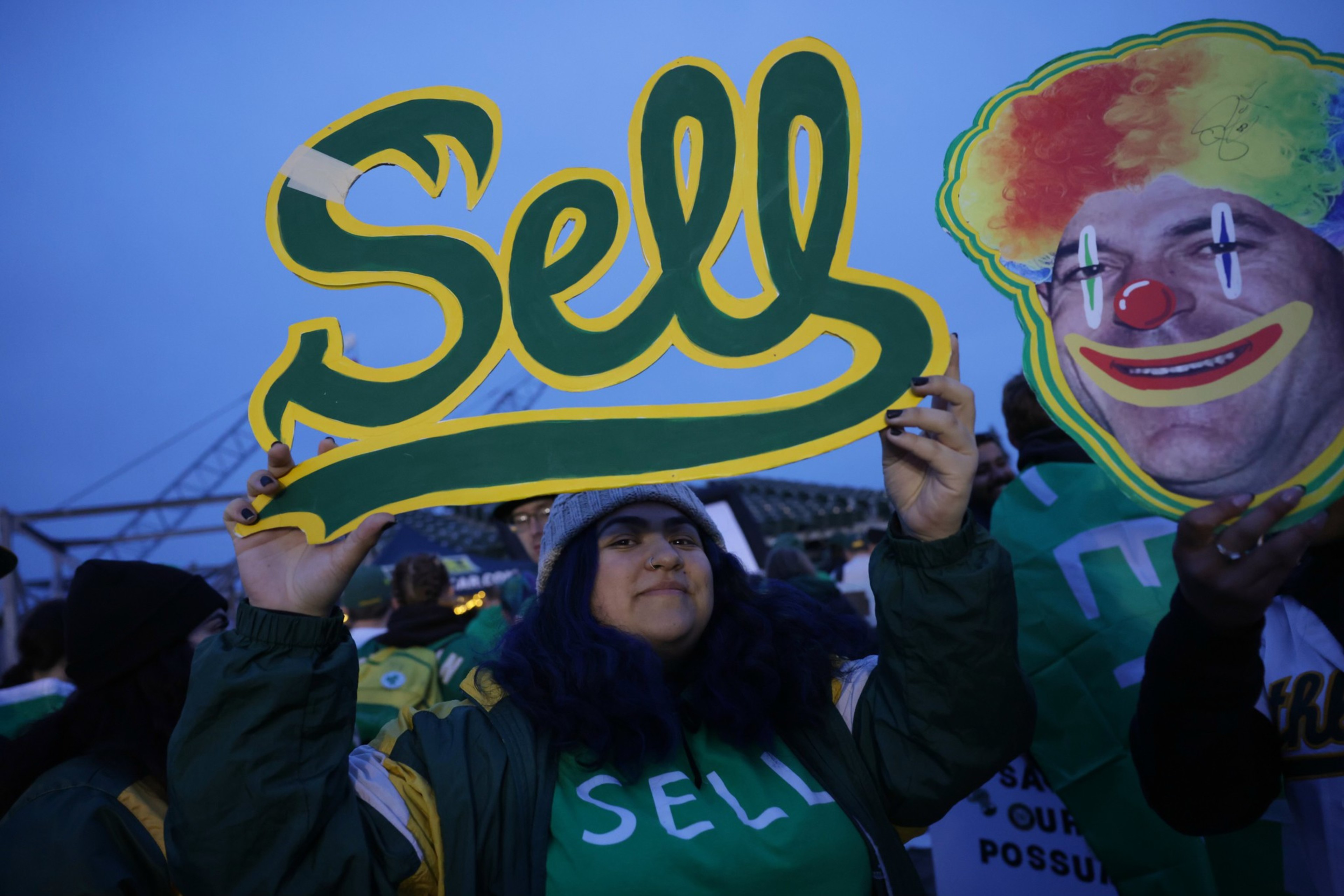 A person is holding up a yellow and green "Sell" sign next to someone with a clown face cutout. They appear to be at an event.