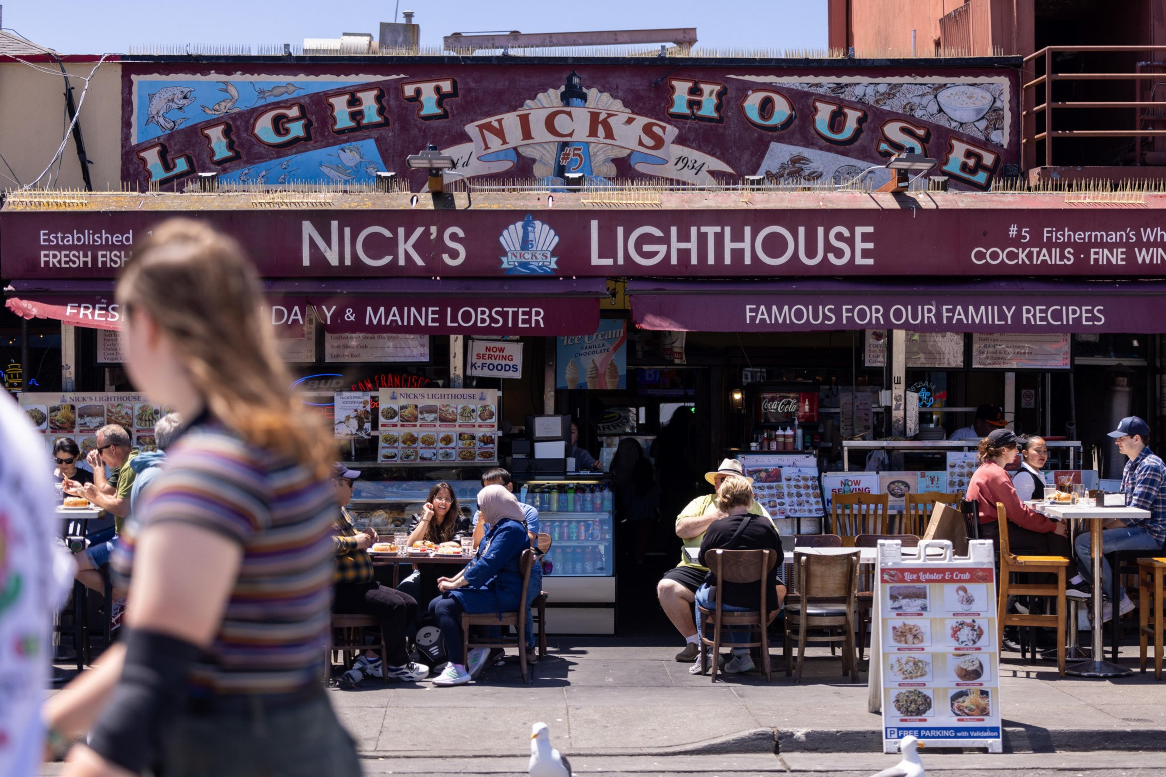 This image shows the exterior of Nick's Lighthouse restaurant, with its maroon canopy displaying its name. People are dining outside on a sunny day, enjoying seafood dishes.