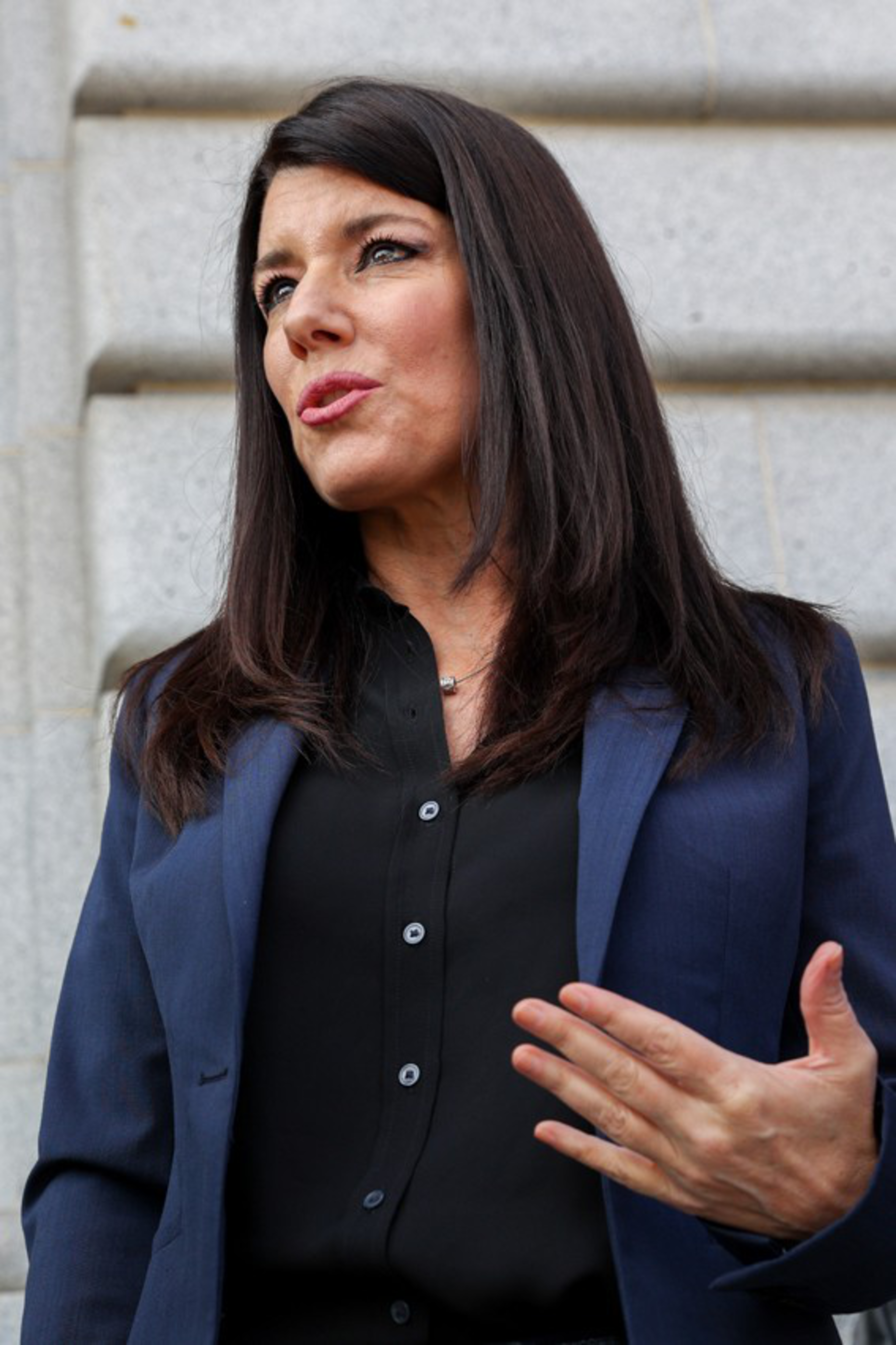 A woman with long dark hair wearing a navy blazer and black shirt gestures with her right hand while speaking, against a gray stone wall background.