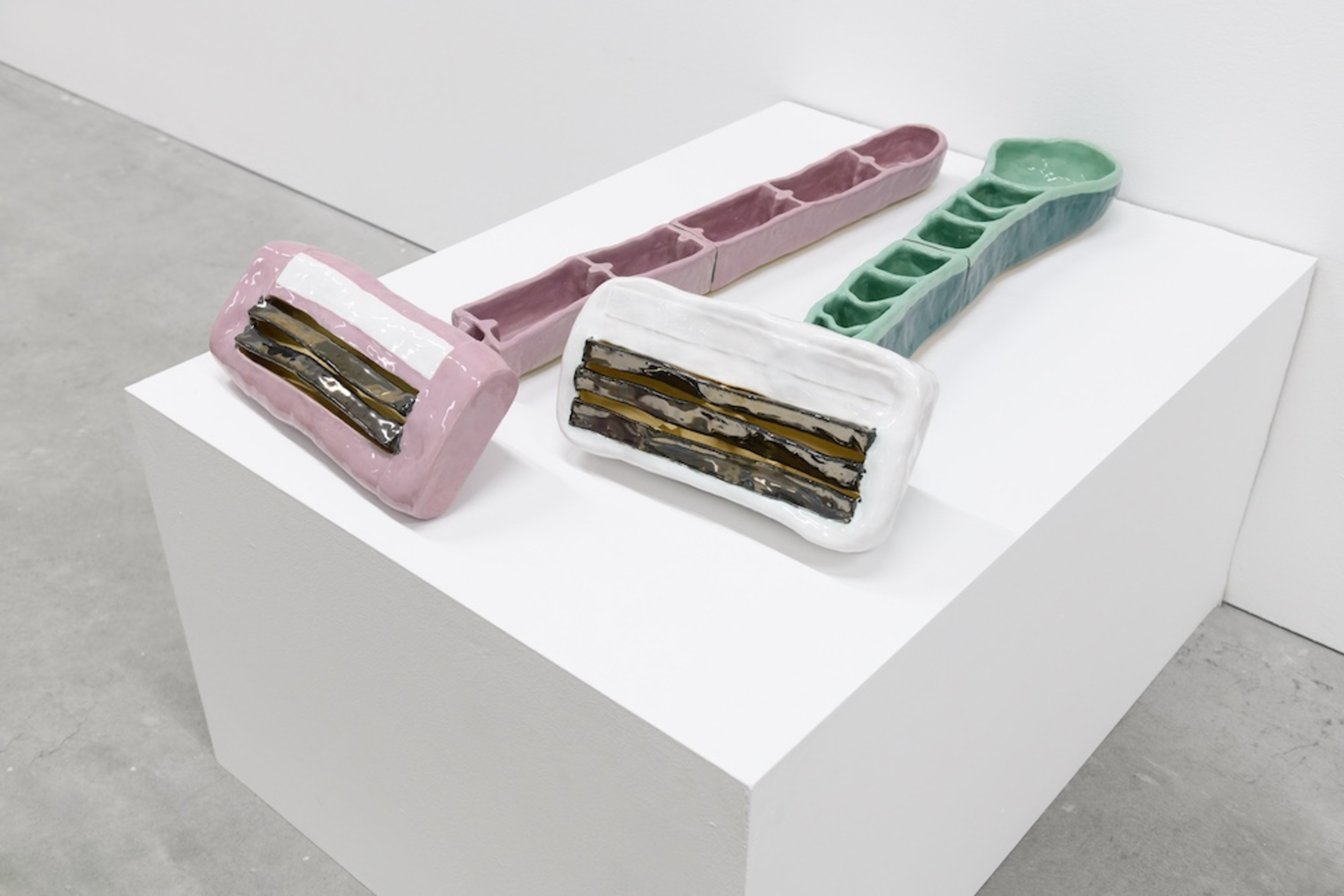 Two oversized ceramic razors with pink and white heads and textured green and pink handles lie on a white pedestal in a gallery space.