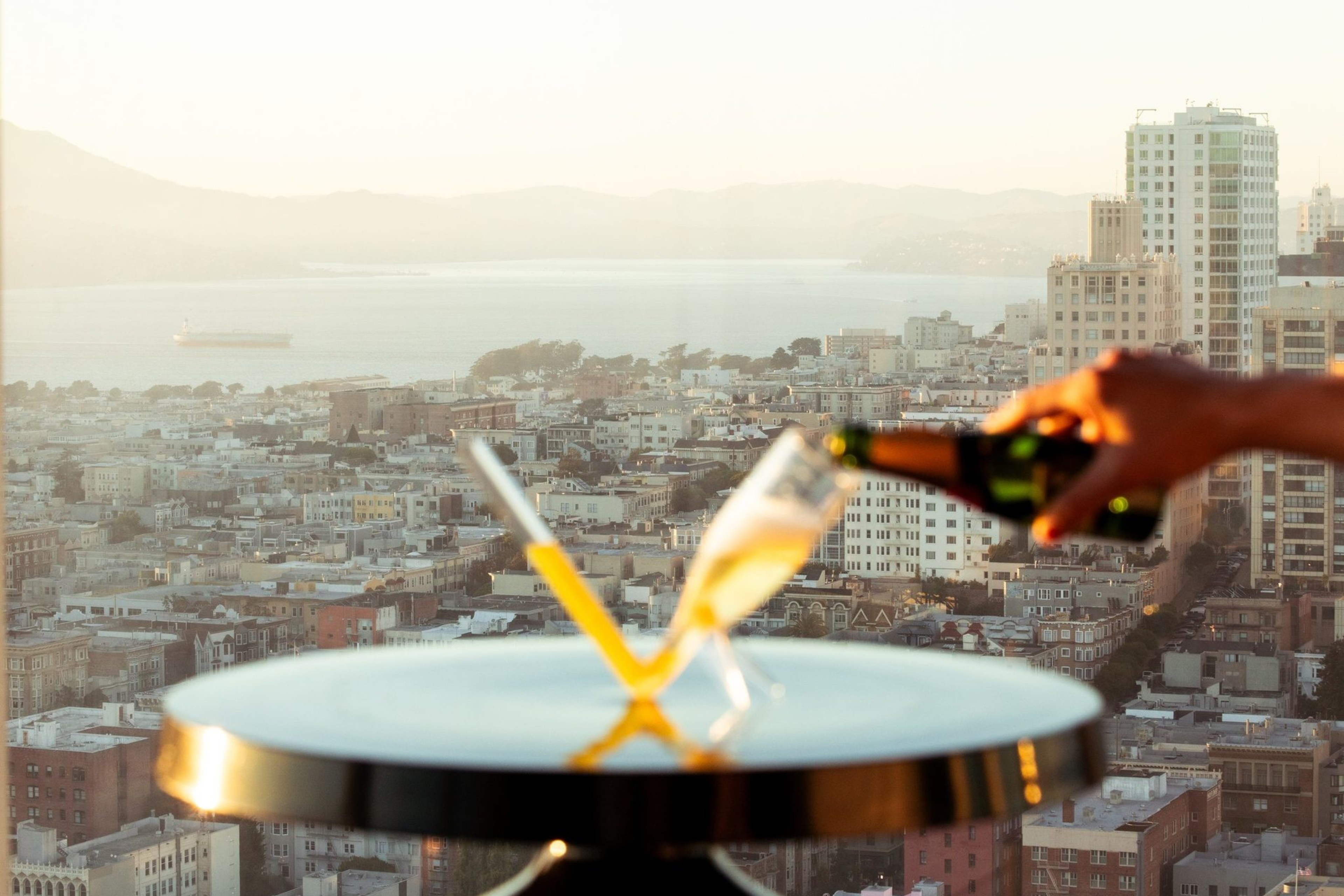 A hand pours champagne into one of two glasses on a small table. In the background, there's a cityscape with buildings, water, and mountains at sunset.