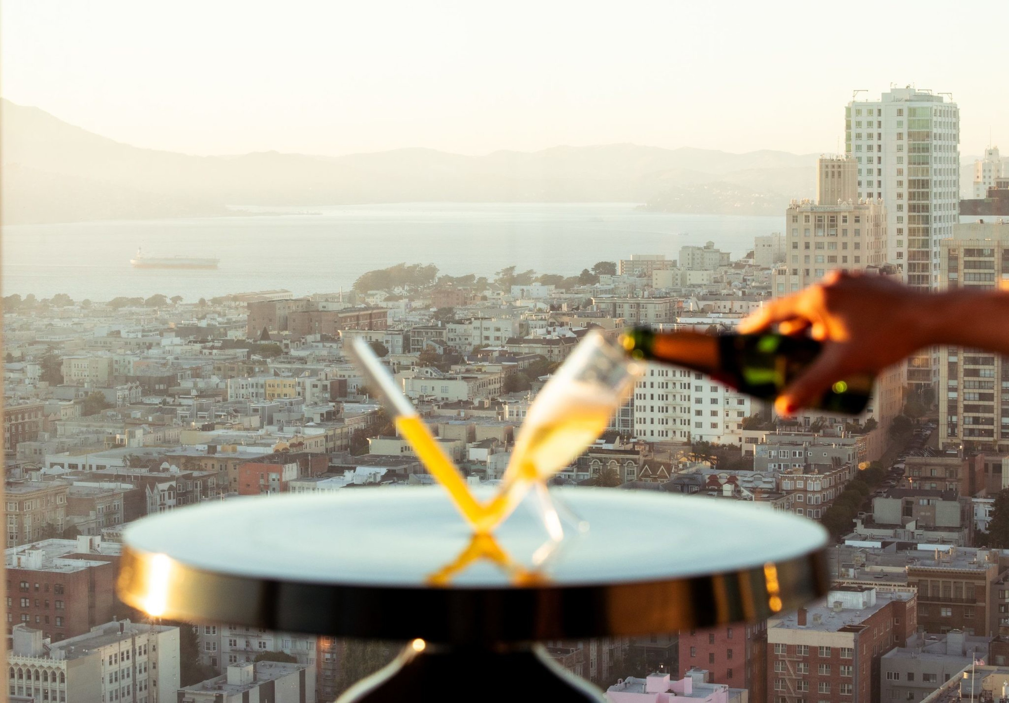 A hand pours champagne into one of two glasses on a small table. In the background, there's a cityscape with buildings, water, and mountains at sunset.