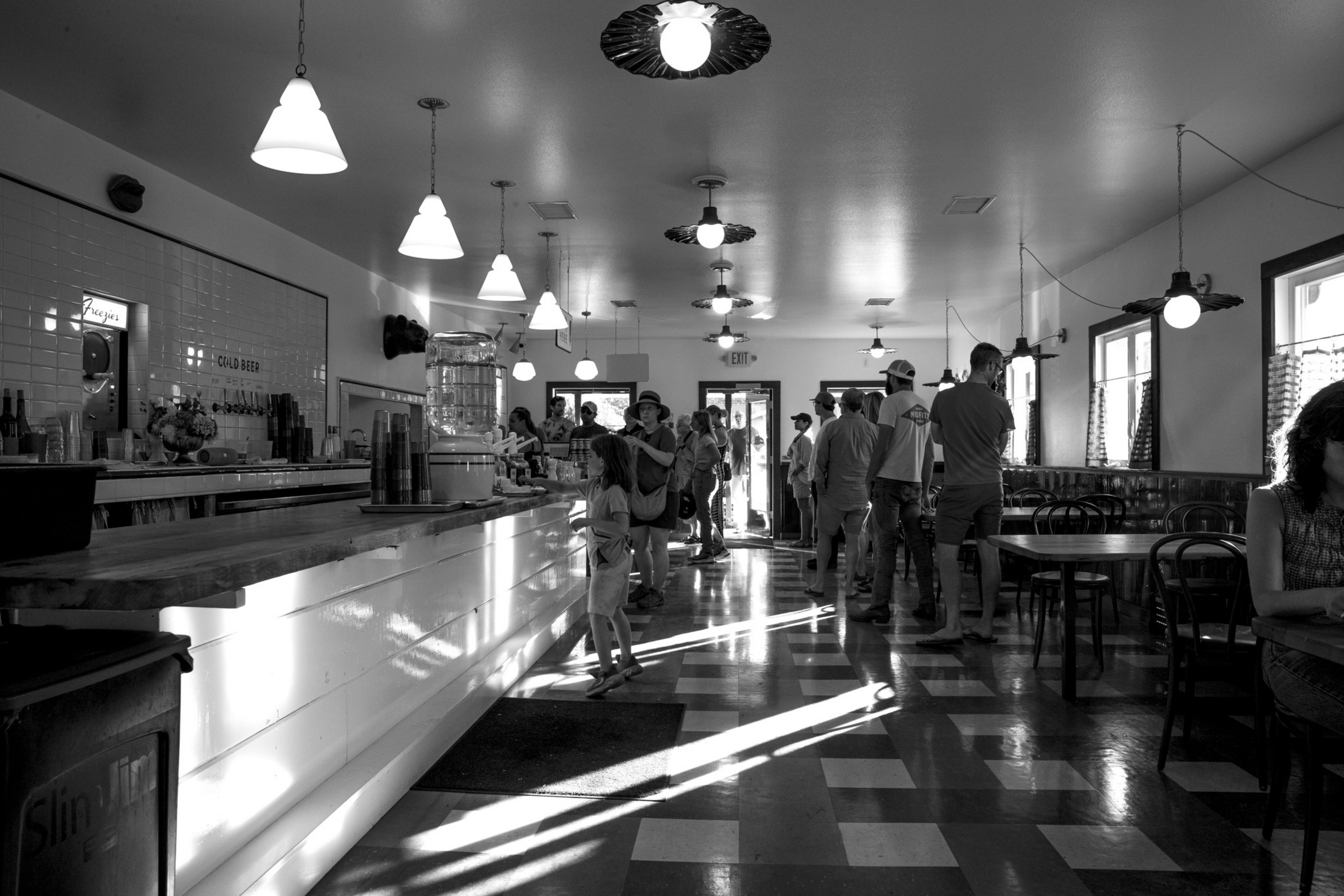 A busy, vintage-style café with checkered floors, hanging lights, and people standing in line by the counter while others sit at tables near windows.