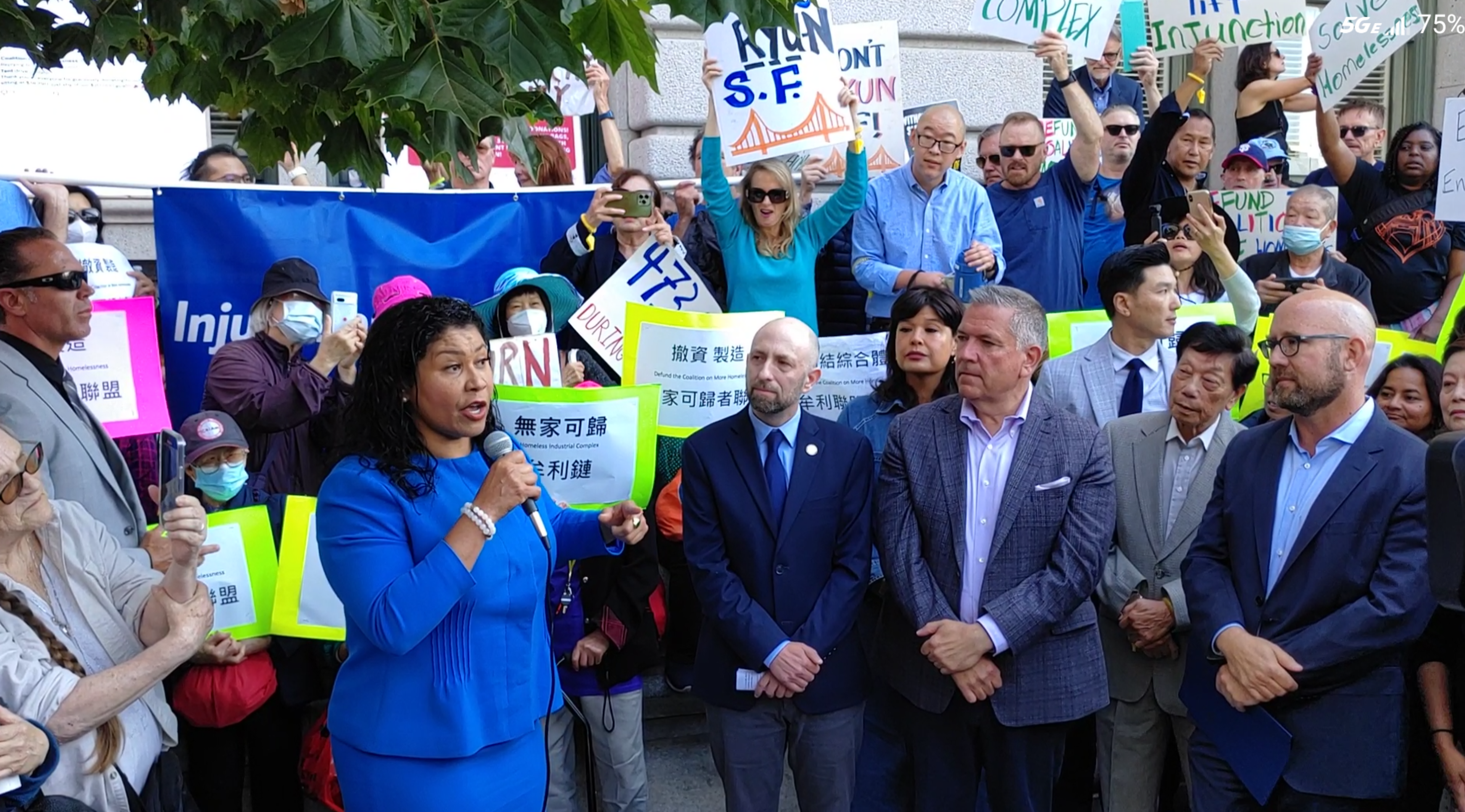 A woman in a blue suit speaks into a microphone, surrounded by men in suits and a crowd holding signs with messages about homelessness and support.