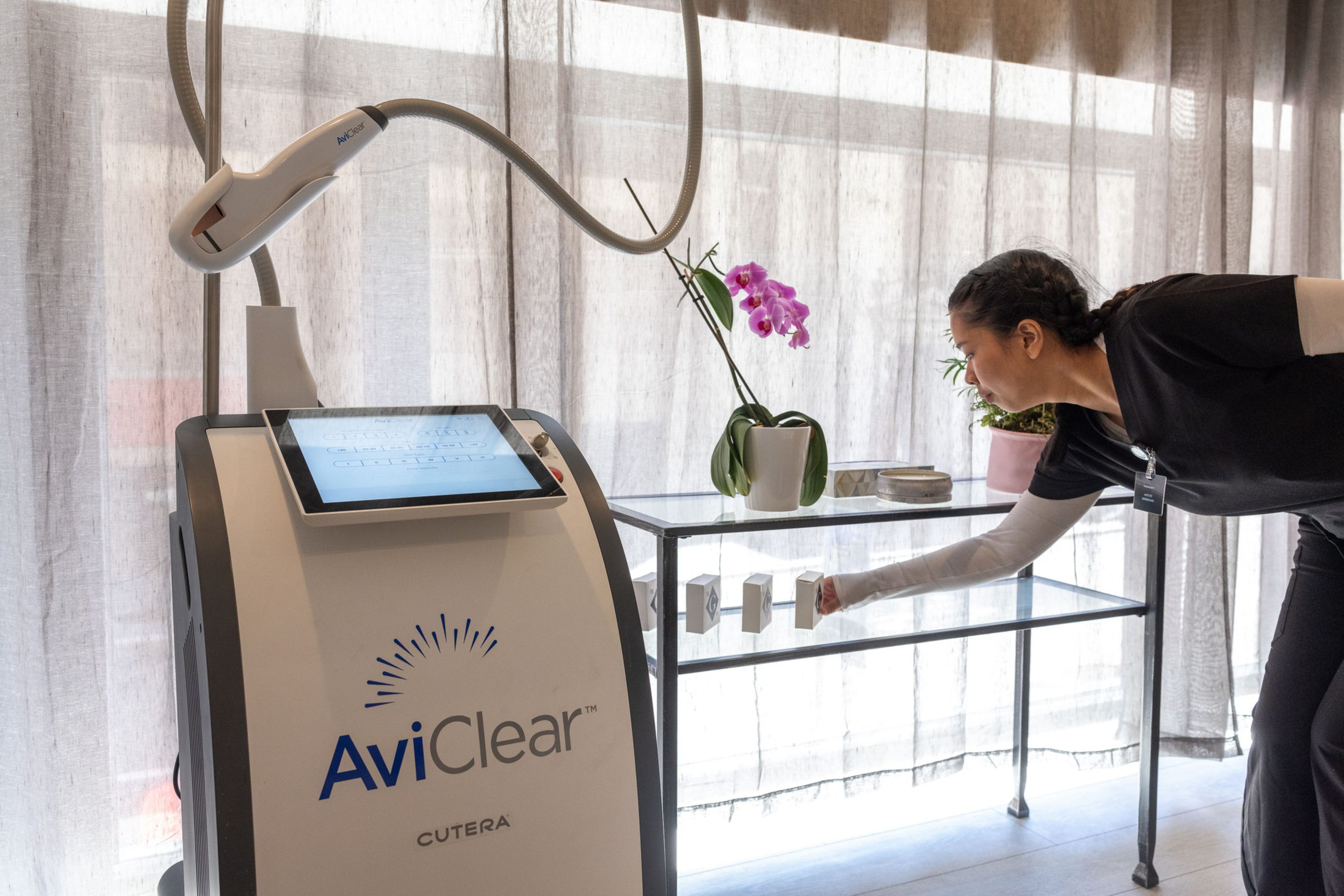 A woman adjusts items on a glass shelf near a large white medical device labeled &quot;AviClear&quot; by Cutera. The scene includes a potted orchid and curtained background.