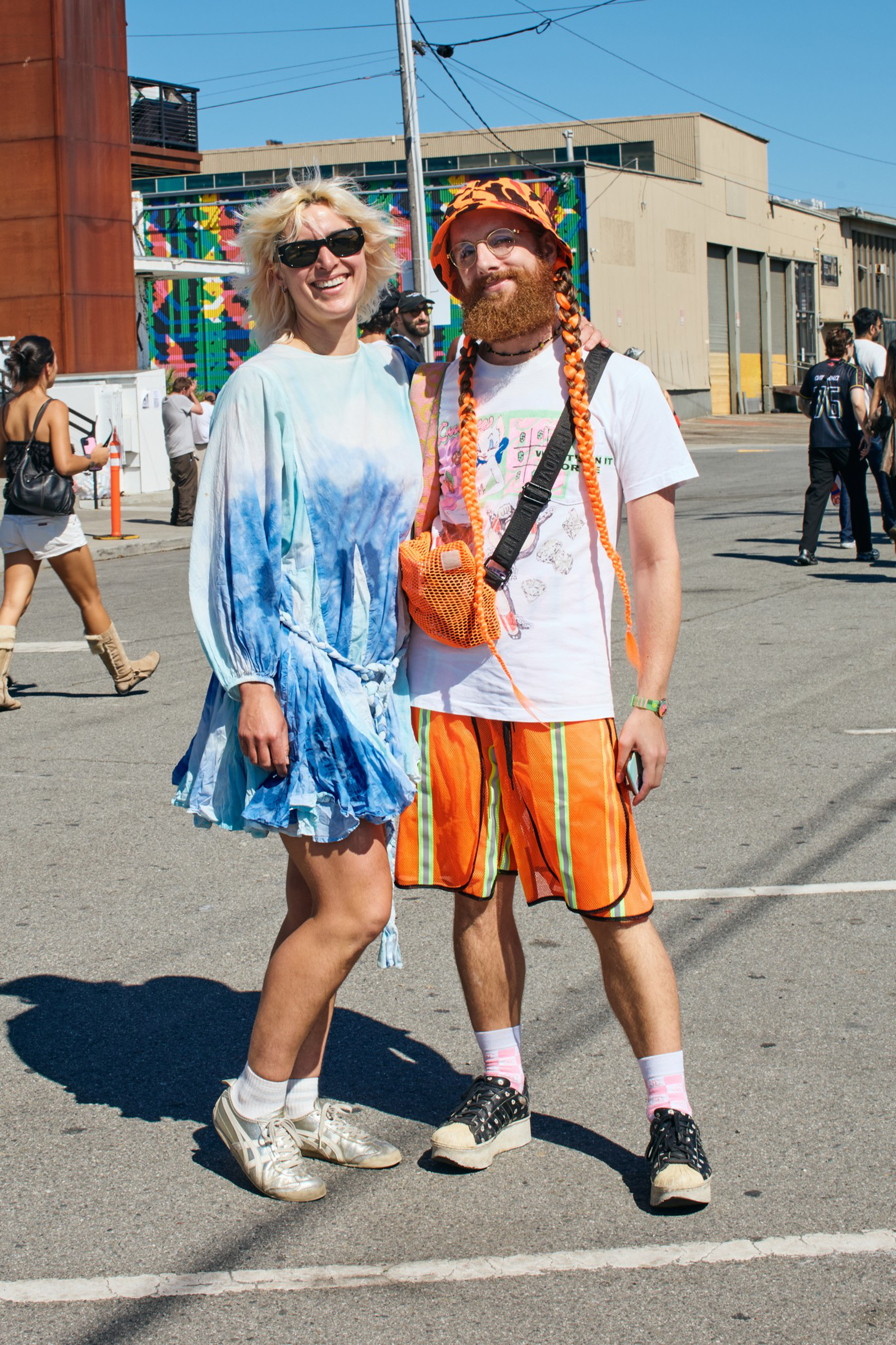Two people stand outdoors on a sunny day, one wearing a blue tie-dye dress and sunglasses, the other in orange-striped shorts, a hat, and braided hair.