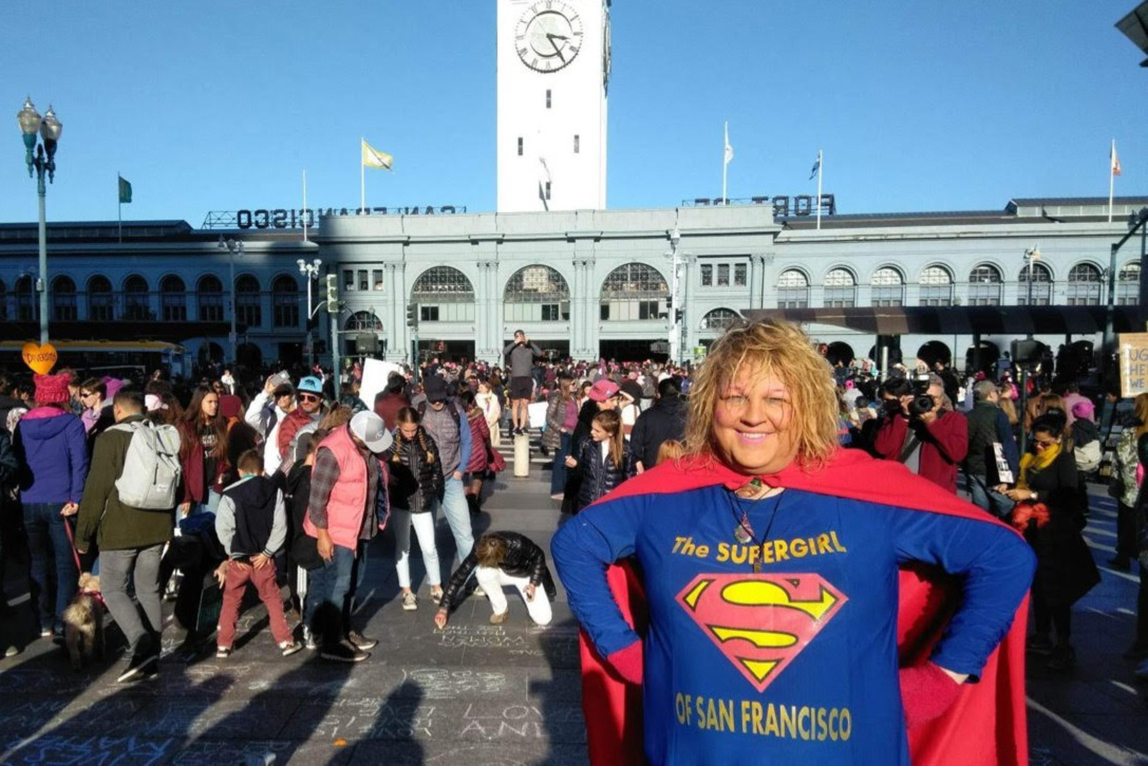 A smiling woman in a "The Supergirl of San Francisco" outfit stands confidently amid a large crowd near a clock tower and historic building.
