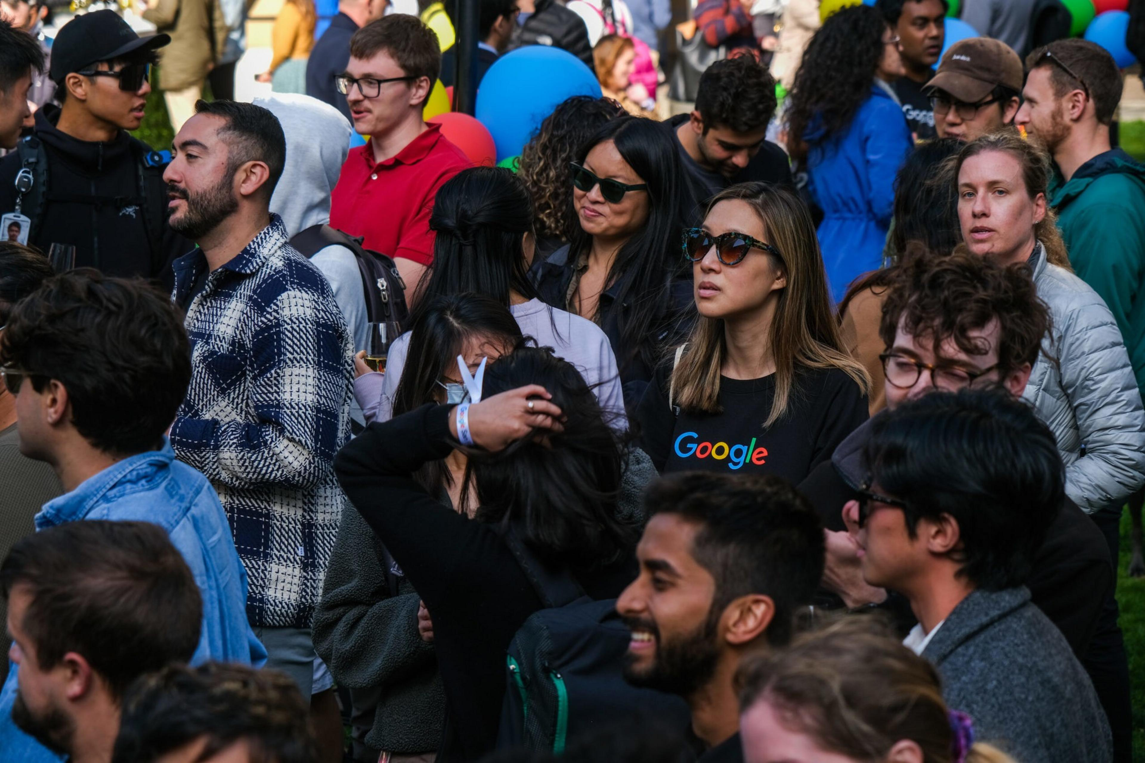 A diverse crowd of people outdoors, some wearing sunglasses, one woman wearing a black shirt with the Google logo, engaging in casual conversation.