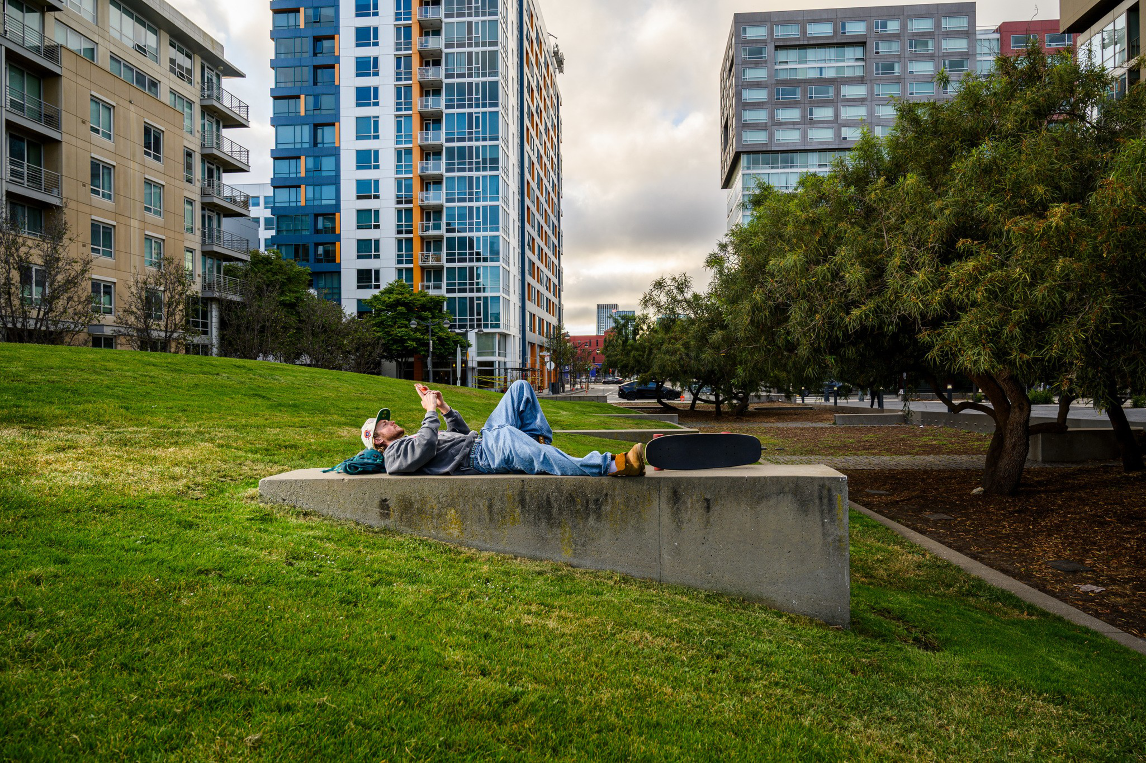 A person lies on a concrete bench in a grassy urban park, holding a phone, with a skateboard resting beside them and buildings in the background.