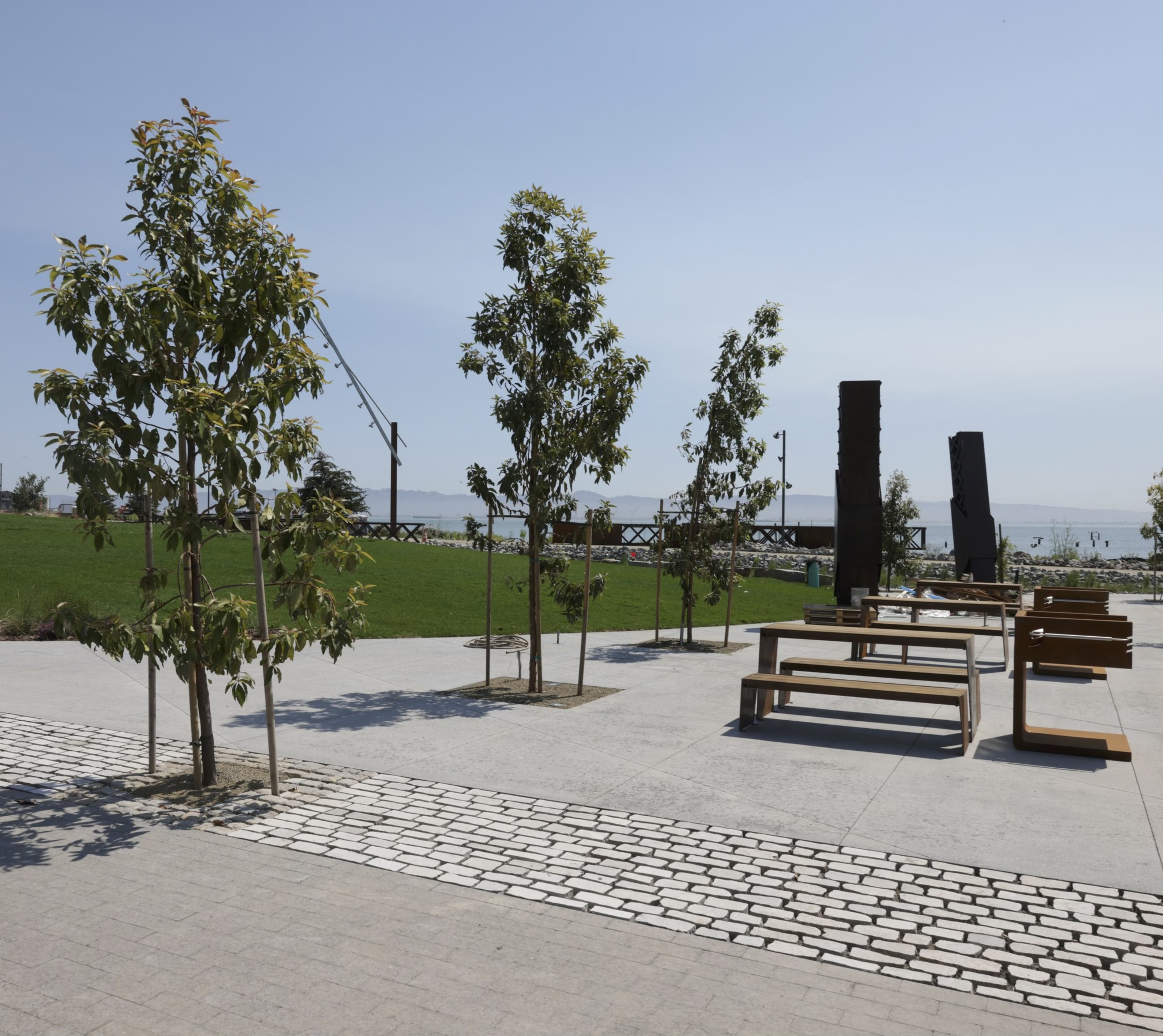 The image shows a park with young trees, modern wooden benches, and a paved walkway beside a grass lawn. In the background, there's a view of distant mountains and a clear sky.