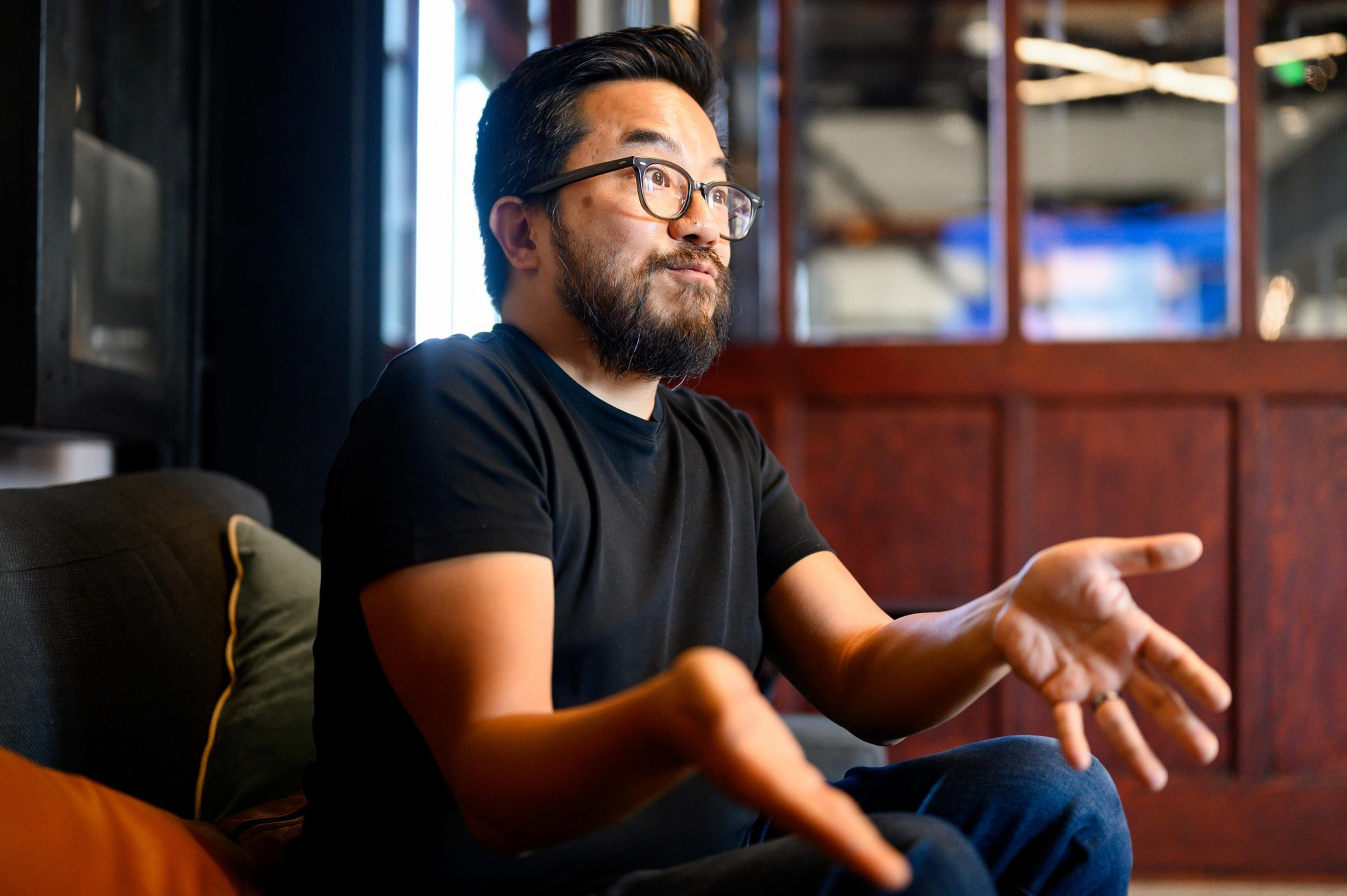 A man with glasses and a beard is sitting on a couch gesturing with his hands. He appears to be explaining something, set against a cozy, blurred background.