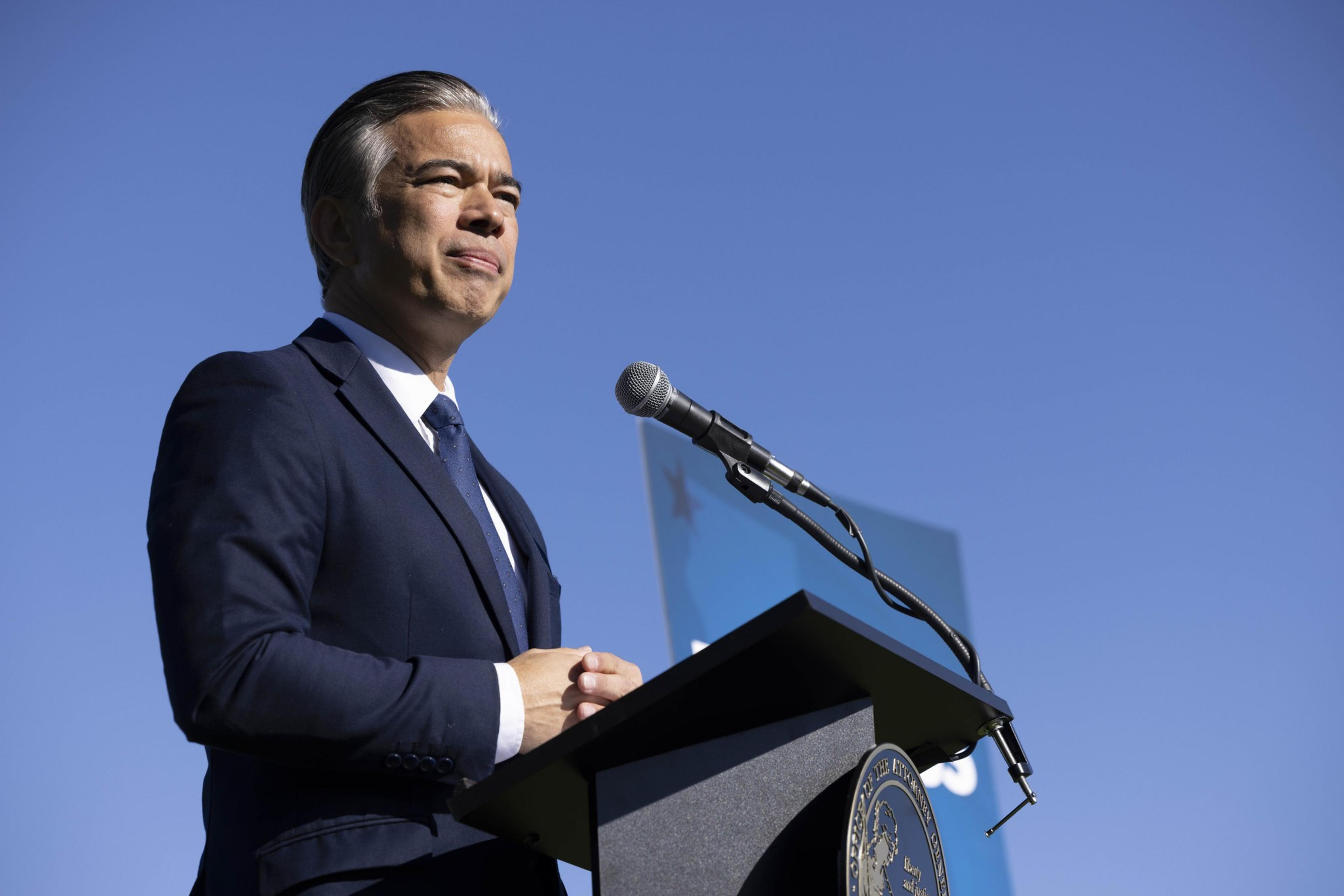 A man in a blue suit stands behind a podium with a microphone, looking thoughtful. The sky is bright blue, and a partial sign is visible in the background.