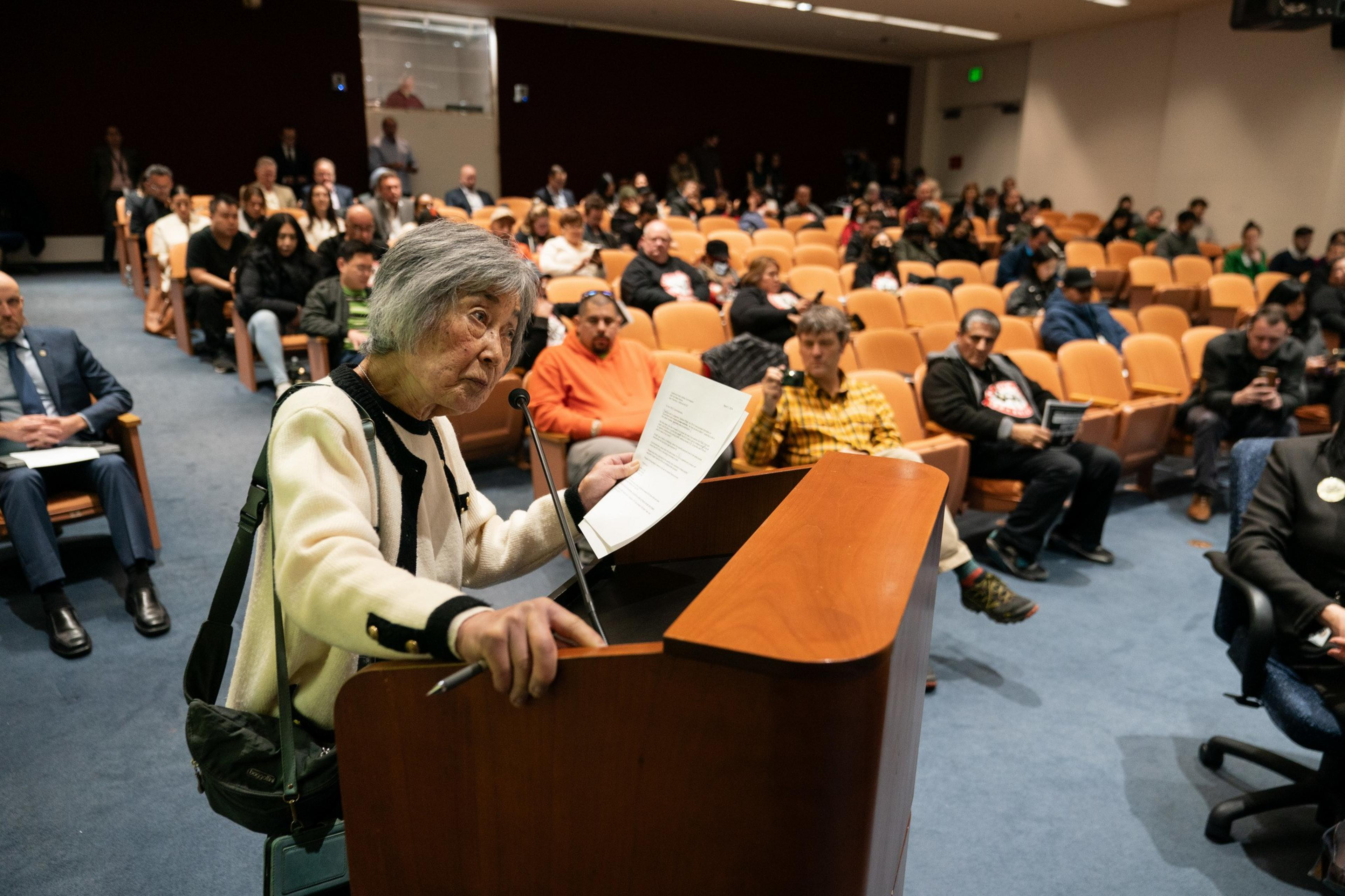 An older person with gray hair speaks at a podium in a hall filled with attendees.