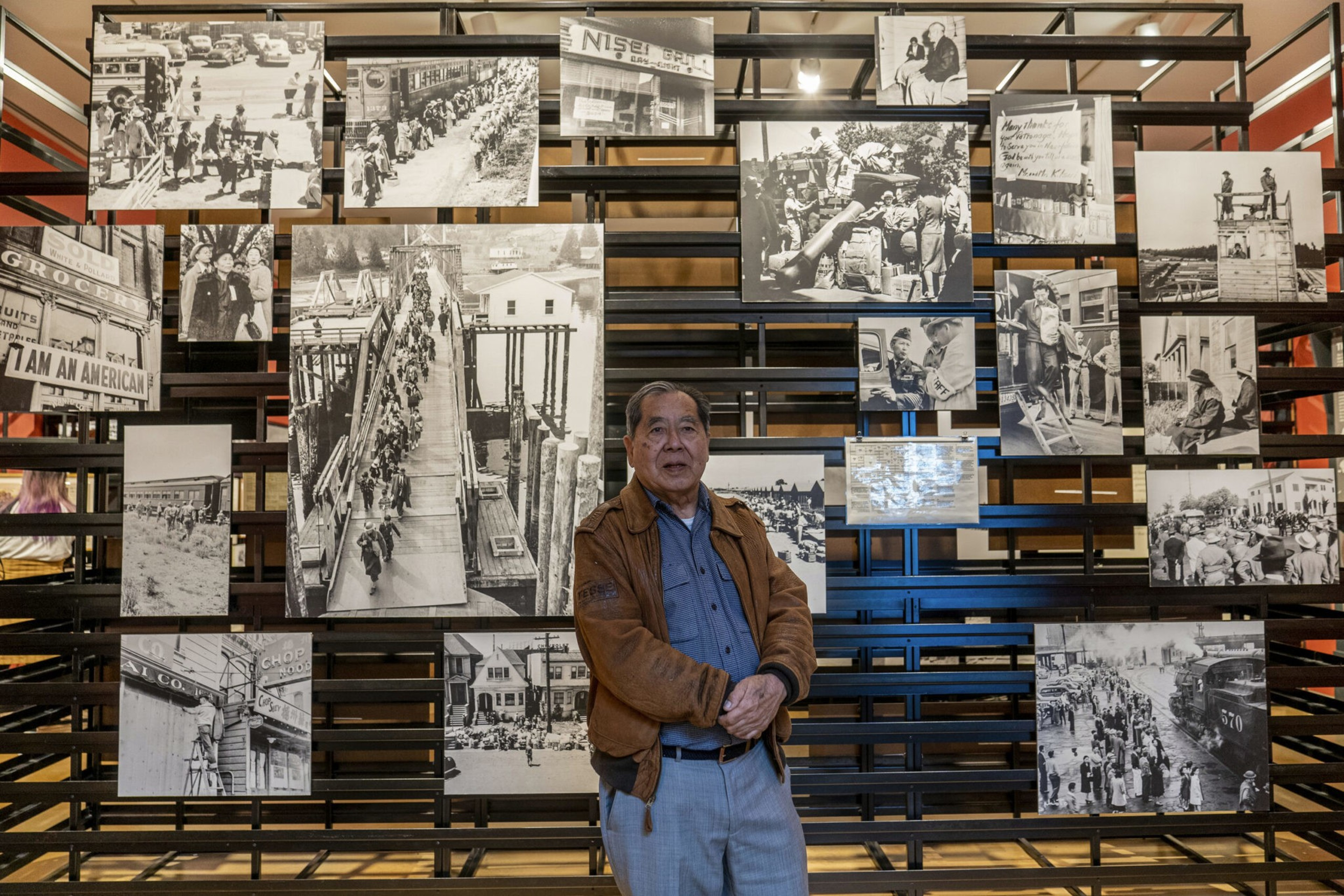 An older man stands in front of a black metal frame displaying numerous historic black-and-white photos depicting crowded streets and community life.
