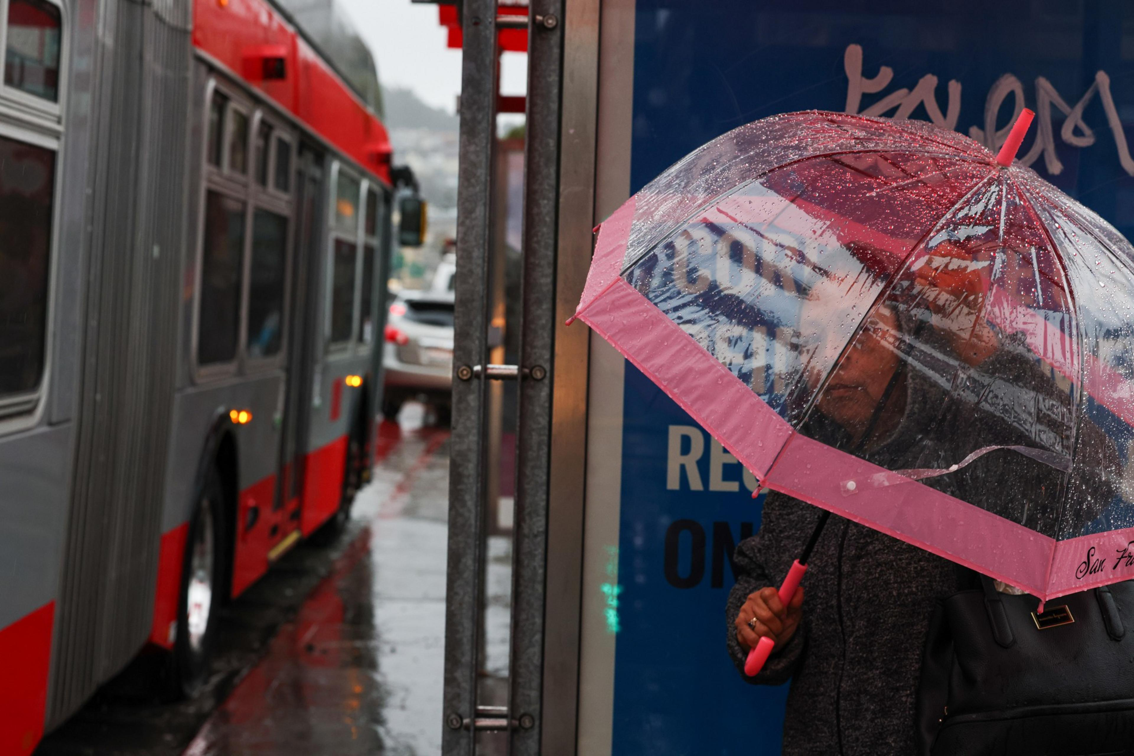 A person stands by a bus shelter on a rainy street, holding a clear umbrella with a pink trim while a red and gray bus passes nearby.