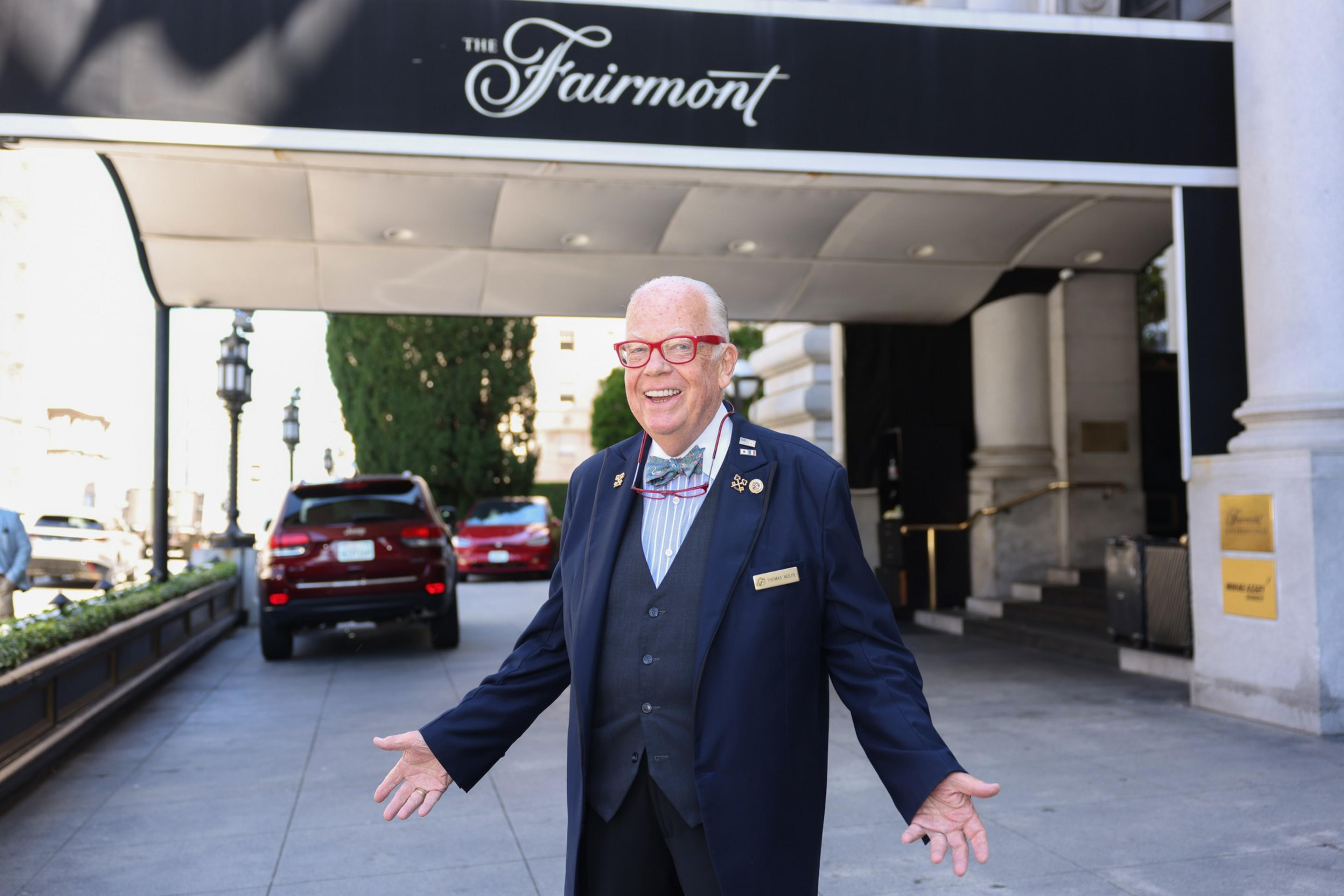 A smiling elderly man in a suit and red glasses stands with open arms under a black canopy labeled "The Fairmont."