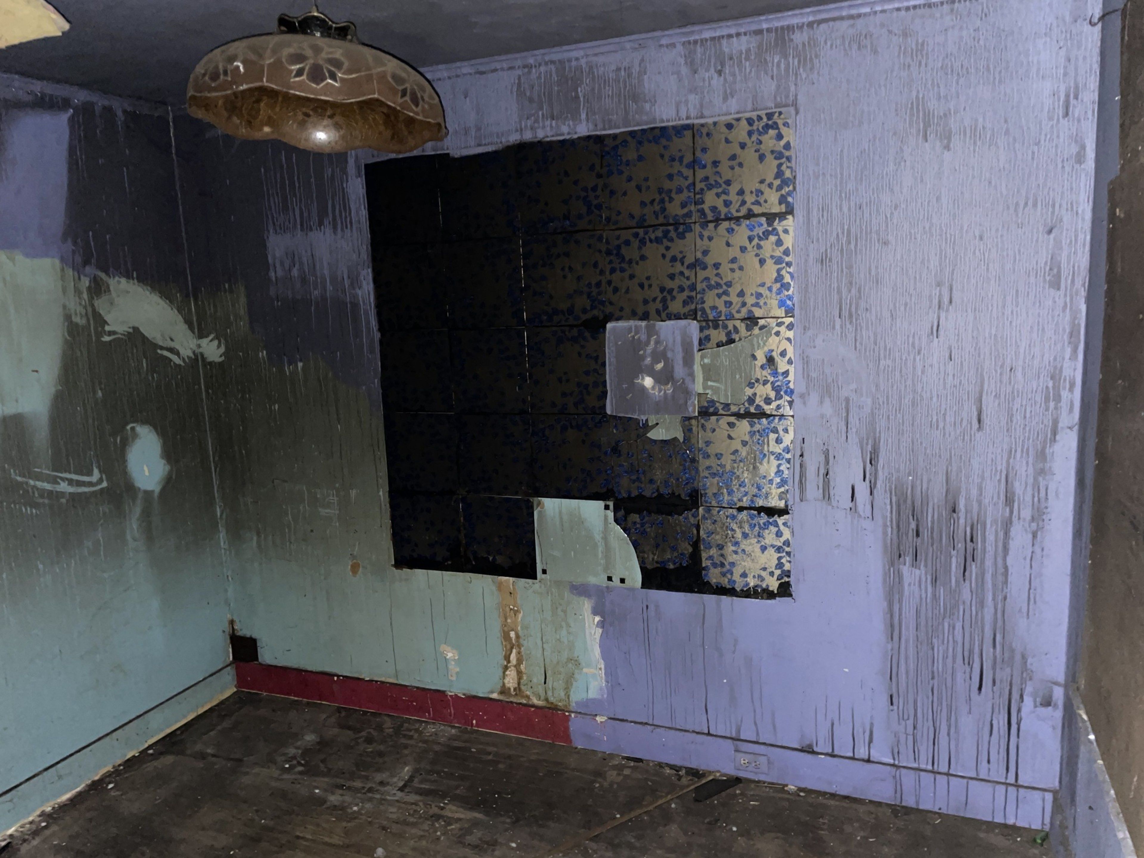 The image shows a dimly lit room with peeling paint on the walls, a broken decorative wall panel, and a vintage light fixture on the ceiling.