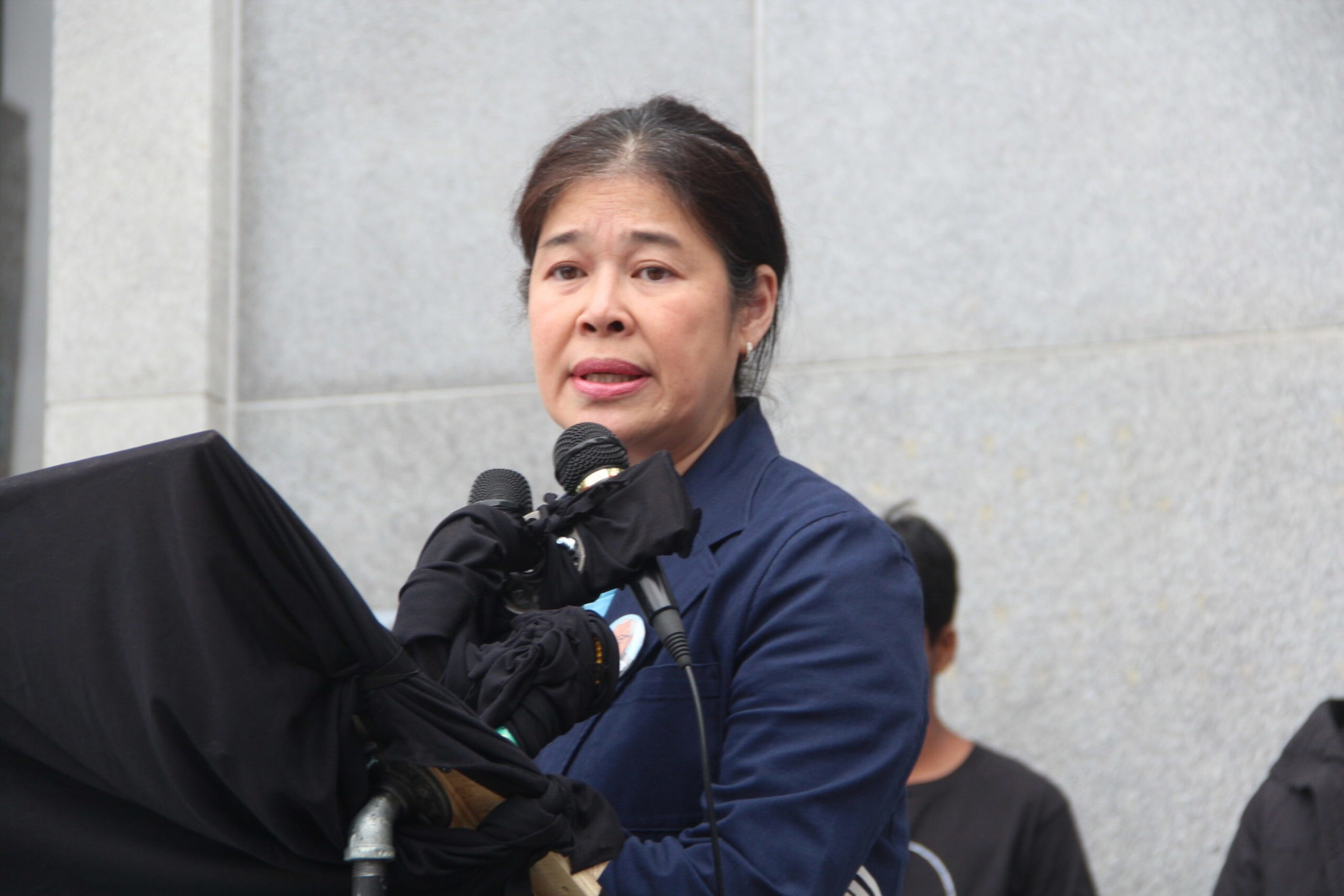 A woman in a navy blazer speaks into two microphones, standing in front of a gray wall with indistinct people blurred in the background.
