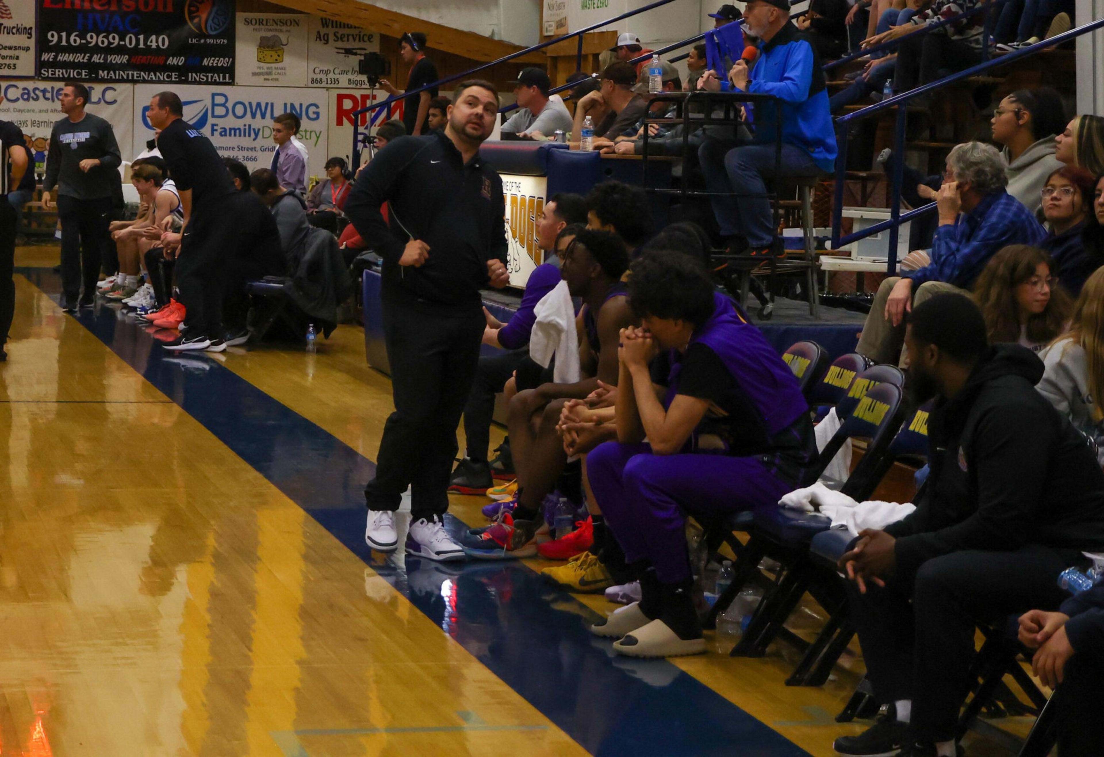 A basketball coach stands near the bench where players in purple uniforms sit, while spectators watch from the bleachers behind them.