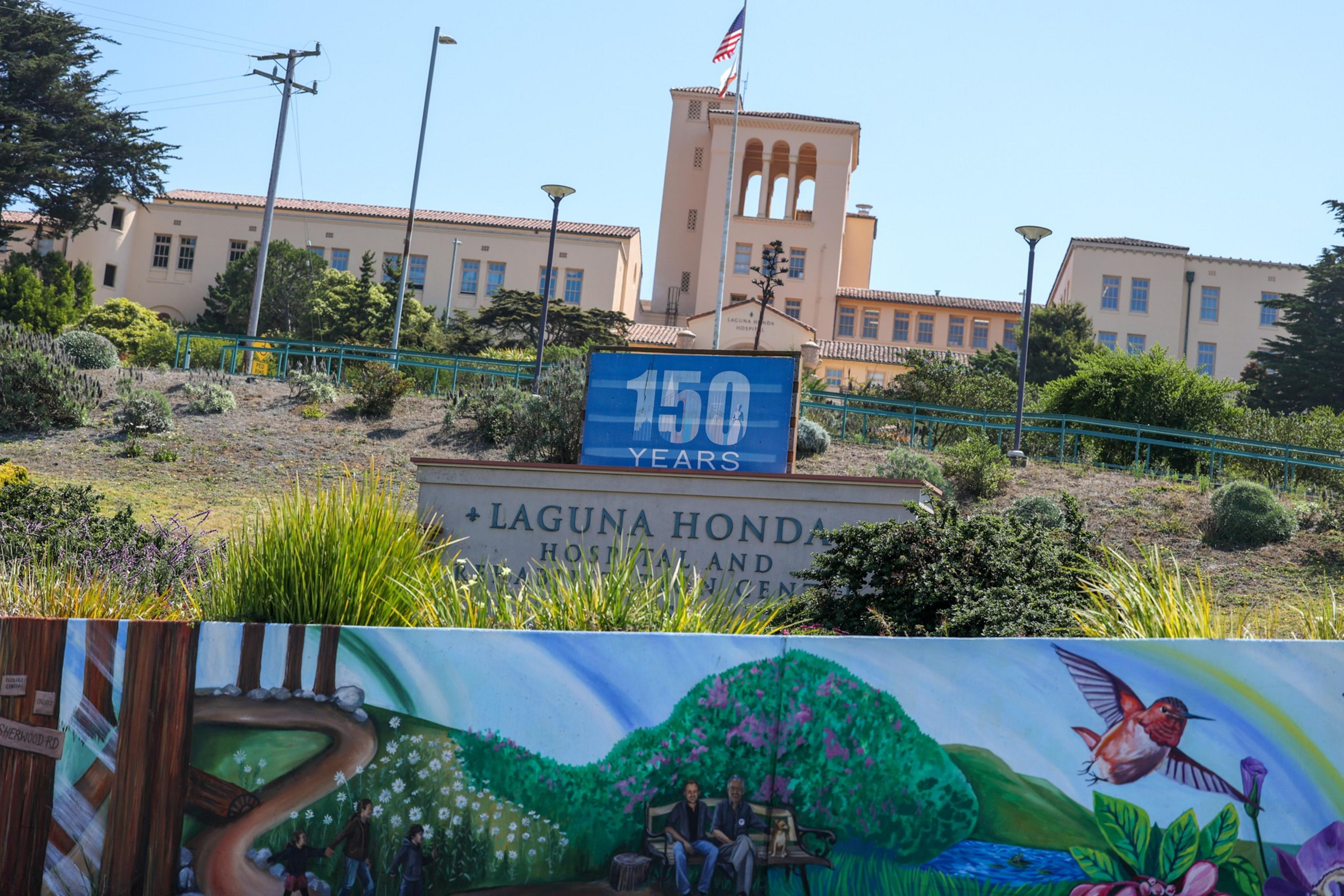 A building with an American flag and a sign reading "150 Years" and "Laguna Honda Hospital and Rehabilitation Center" sits behind a colorful mural of people and nature.