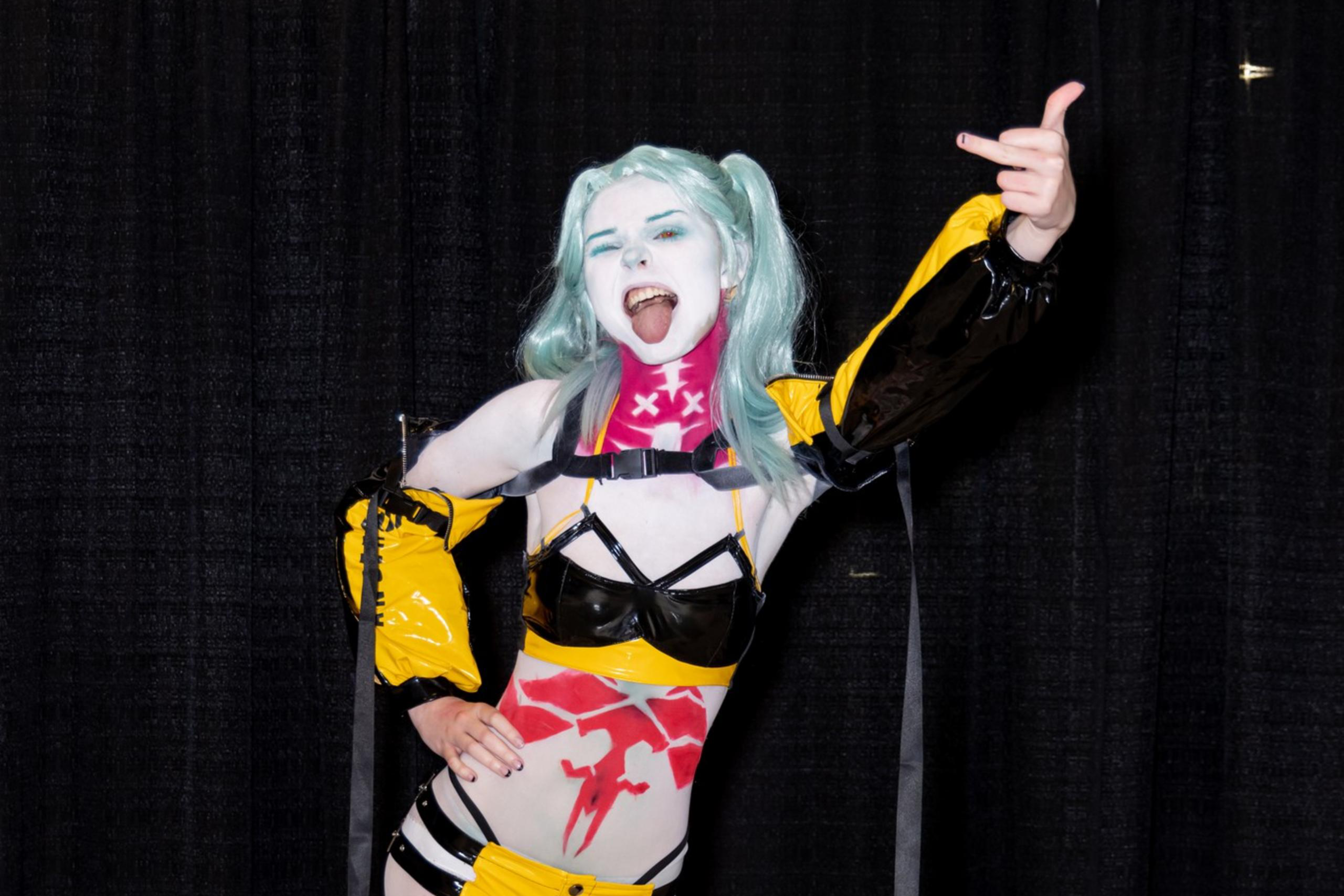 A person in bold cosplay with light blue hair, expressive makeup, and an outfit combining black, yellow, and red elements poses energetically against a black curtain.