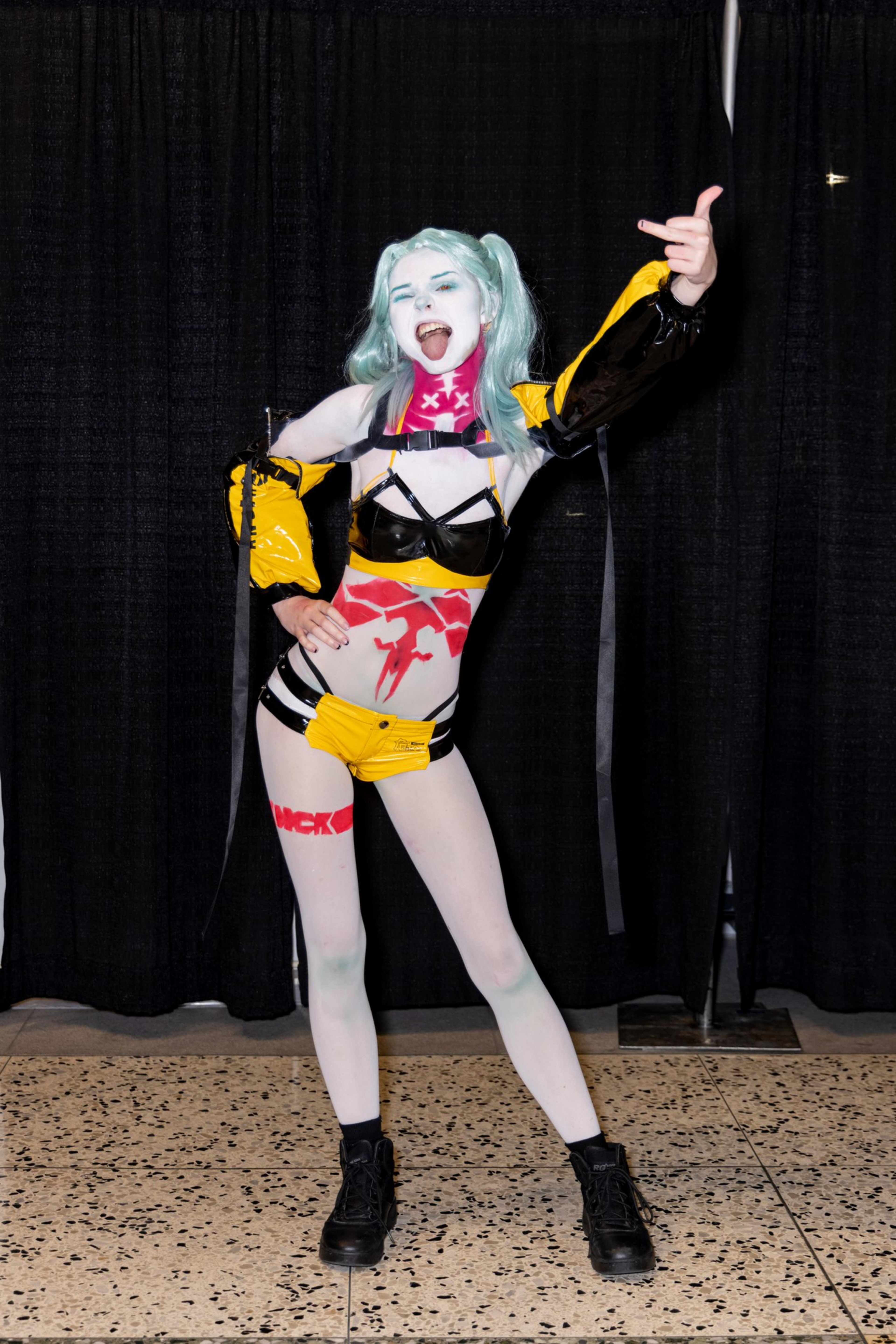 A person in bold cosplay with light blue hair, expressive makeup, and an outfit combining black, yellow, and red elements poses energetically against a black curtain.
