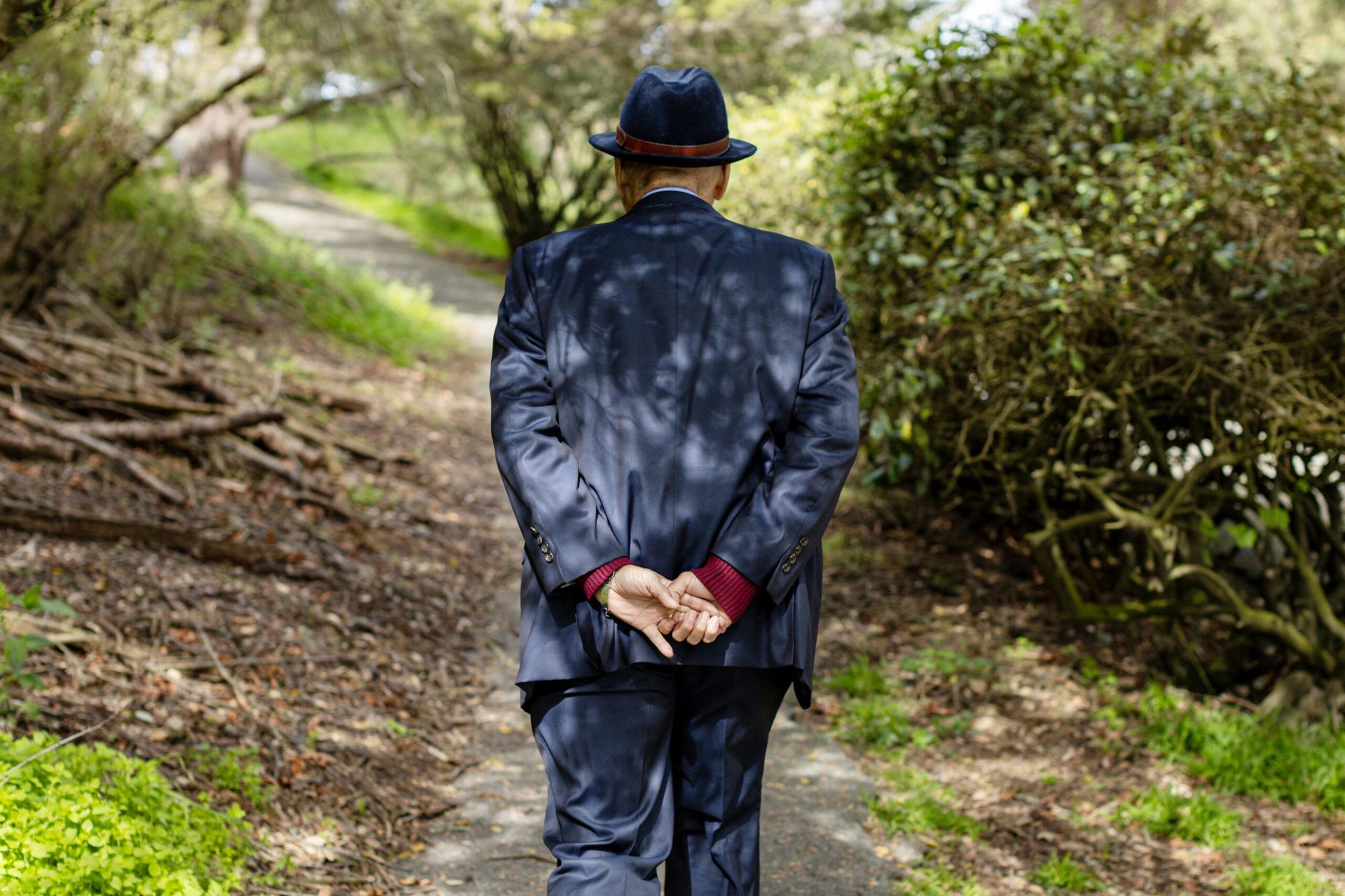 An elderly man in a navy blue suit and hat walks with his hands clasped behind his back on a shaded, winding forest path.